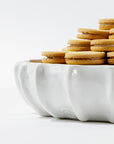 Bowl No. 727 by Montes Doggett, a white, high-fired ceramic piece handcrafted in Peru, is elegantly filled with a stack of cream-filled sandwich cookies arranged neatly. The bowl features a wavy, scalloped edge design that stands out against the plain white background, drawing attention to both the handmade ceramic bowl and the delicious cookies.