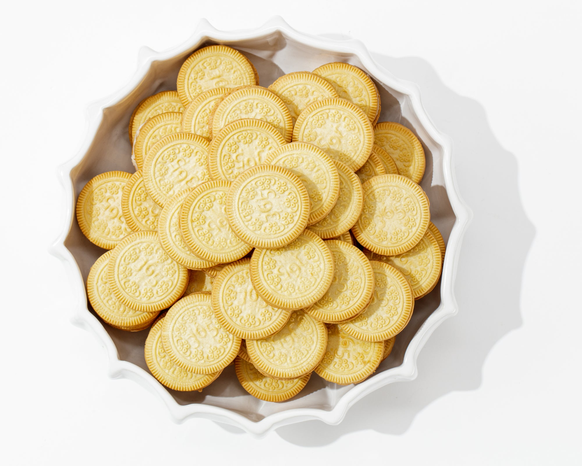 Bowl No. 727 from Montes Doggett, a white, scalloped-edge handmade ceramic creation filled with golden sandwich cookies with cream filling. The cookies feature intricate embossed designs on the top, and the bowl, crafted by skilled artisans in Peru using fine quality clays, is set against a white background.