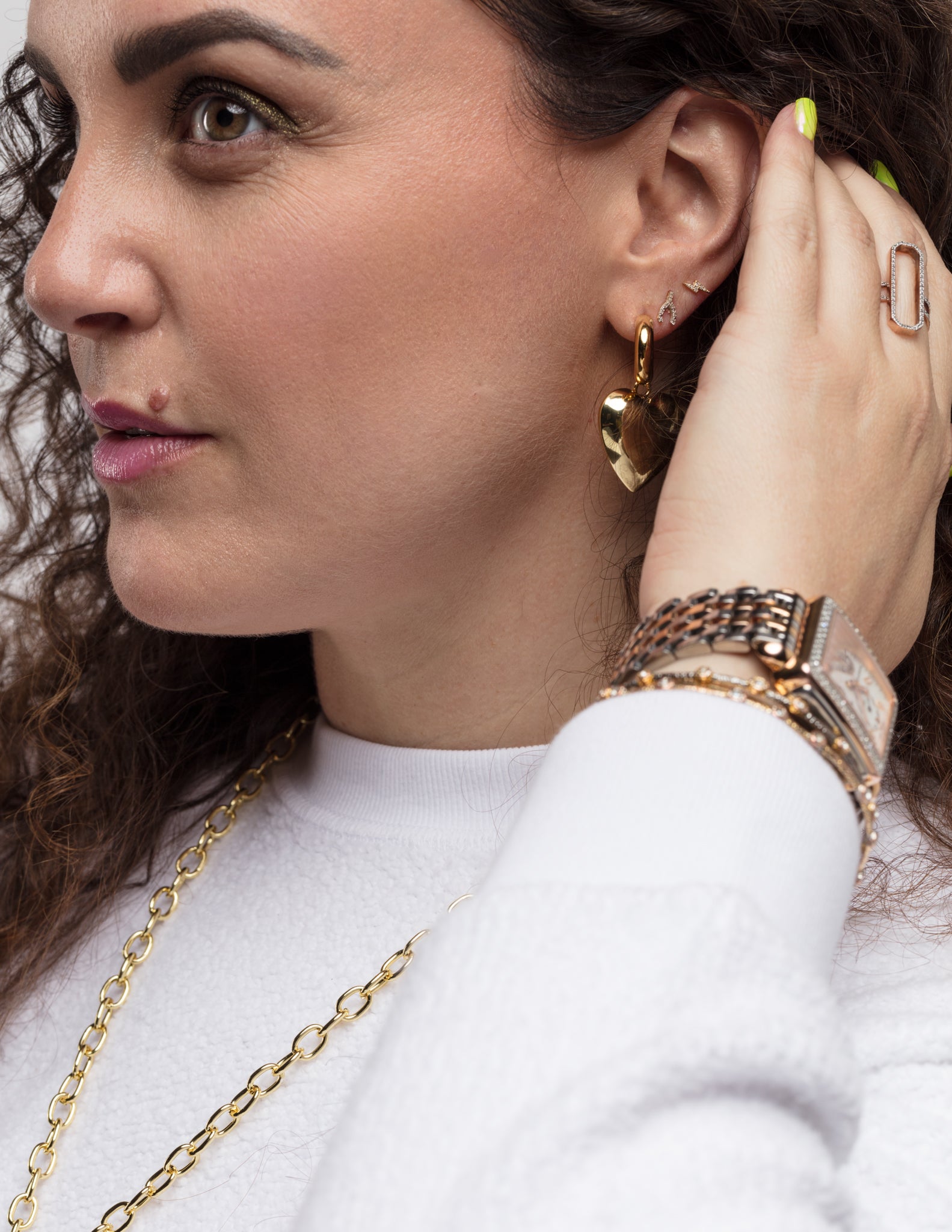 Close-up of a person with curly hair wearing several pieces of jewelry: Bubble Heart Earrings by Lucky Star, a pendant necklace with thick links, multiple rings, and bracelets with a watch. This person has a thoughtful expression and is touching their ear with one hand.