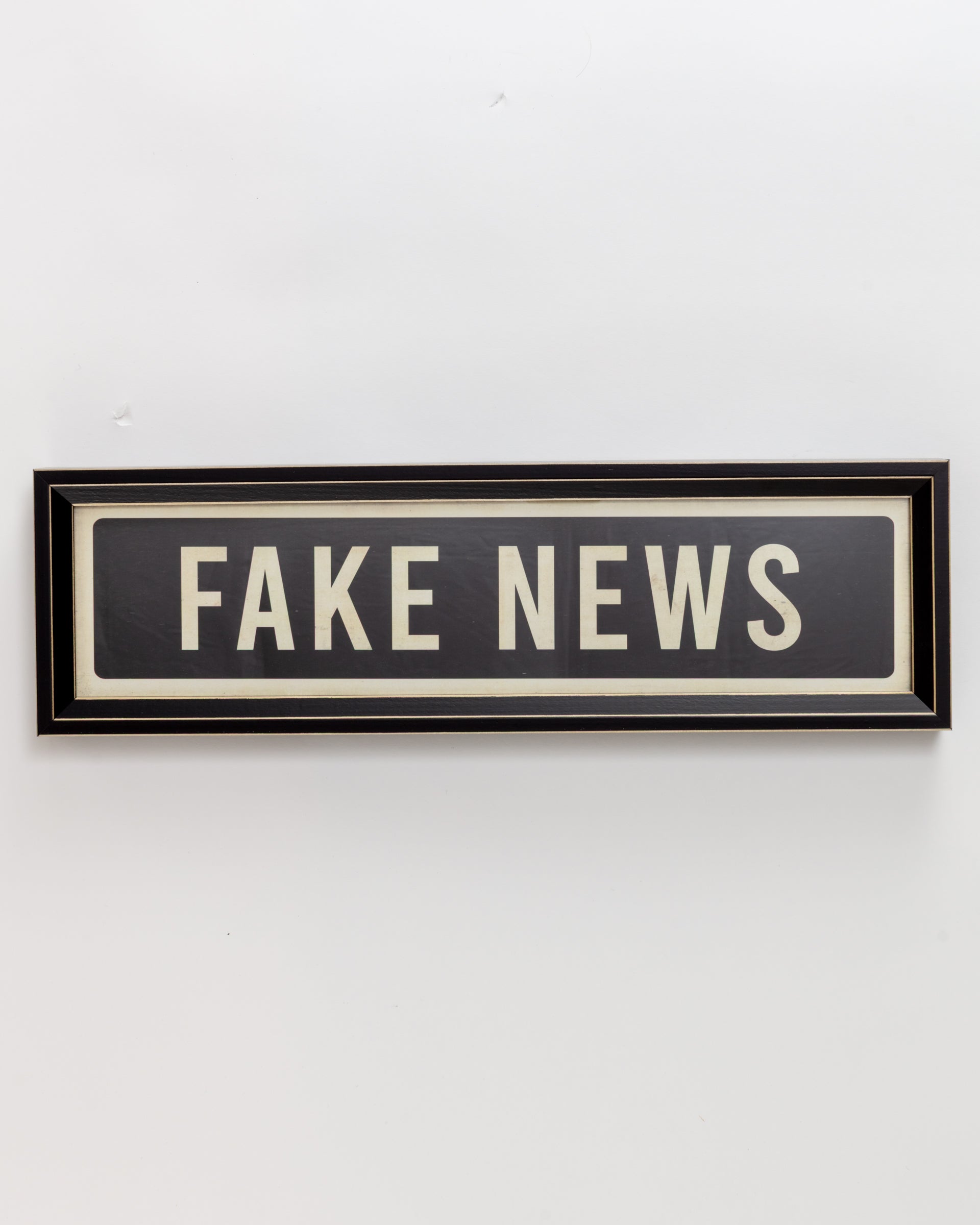 A framed "Small Sign In Black" by Spicher And Company, featuring bold white letters spelling "FAKE NEWS" on a black background, hangs against a plain white wall, reminiscent of a custom skateboard deck design.