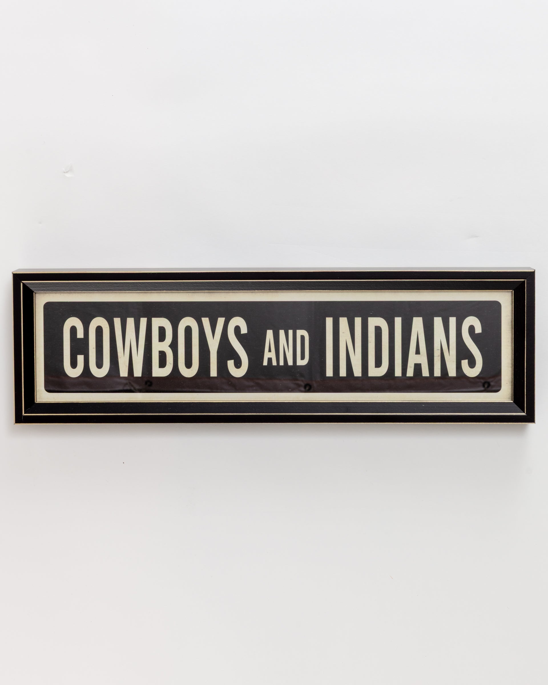 The "Small Sign In Black" by Spicher And Company features a black background and white text, reading "COWBOYS AND INDIANS." Its sleek, narrow frame offers a simple, vintage appeal akin to a custom skateboard deck.