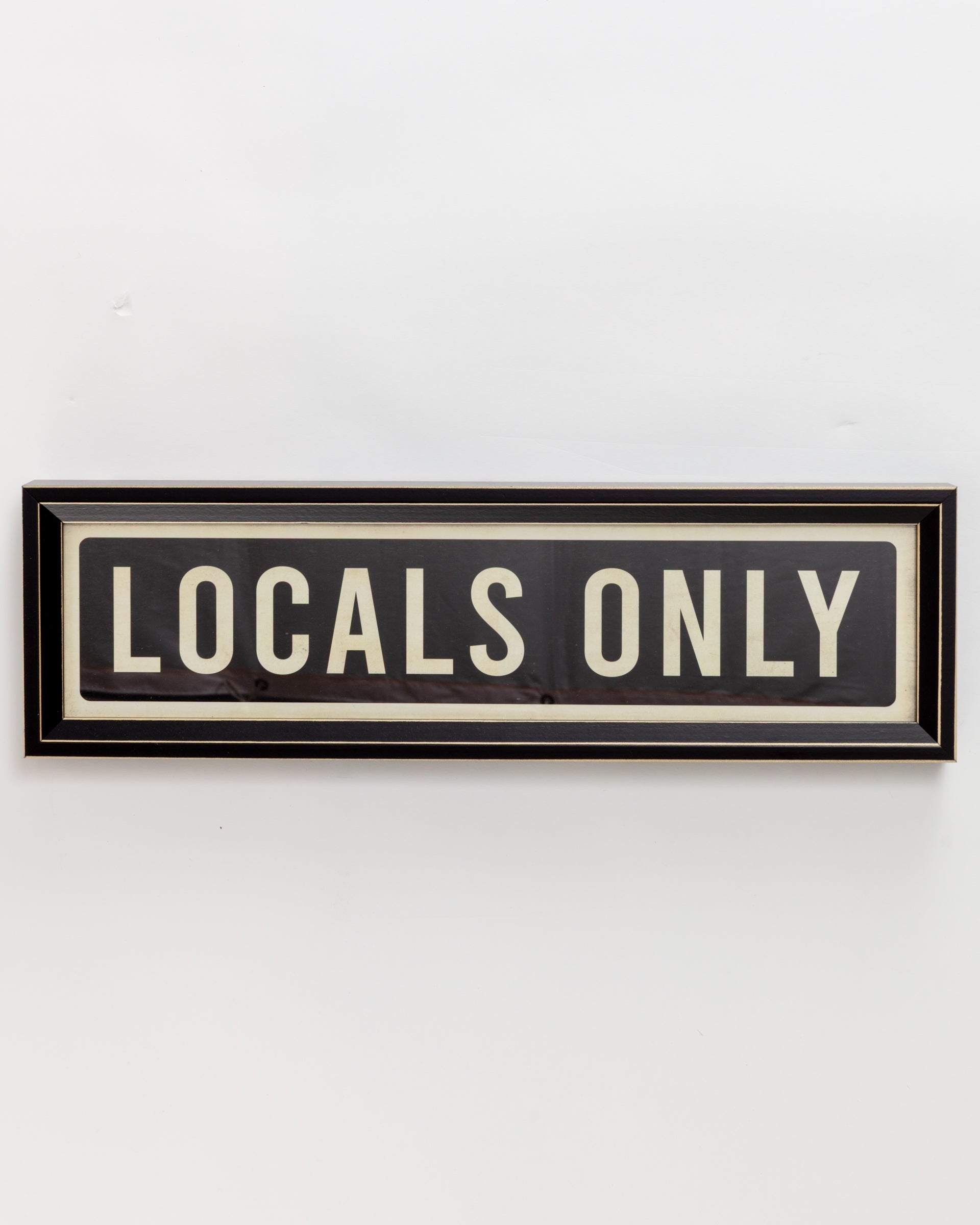 A black-framed rectangular sign from Spicher And Company, "Small Sign In Black," features bold white lettering that reads "LOCALS ONLY" against a plain white wall, similar to a custom skateboard deck's striking design.