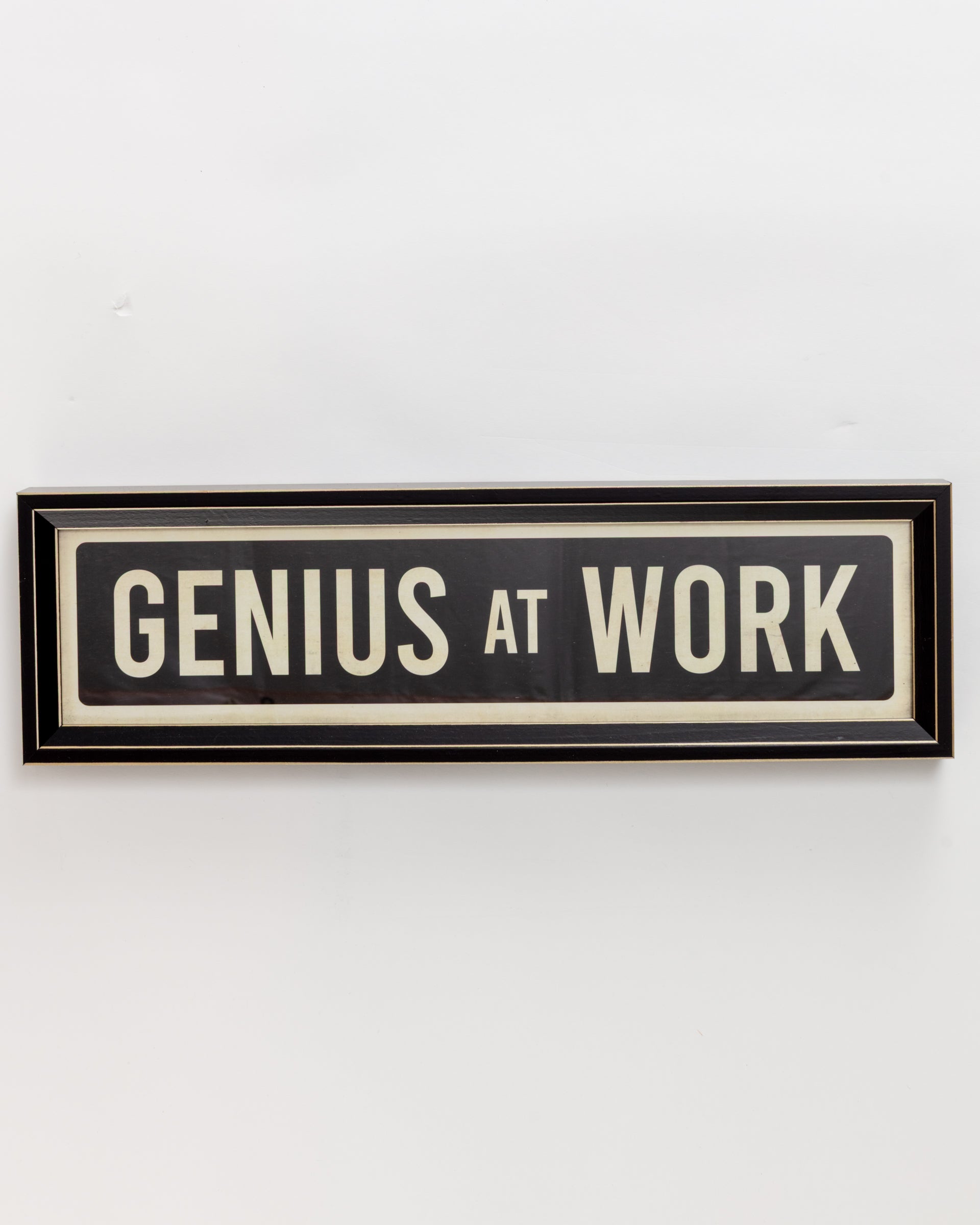 The "Small Sign In Black" by Spicher And Company features a black rectangular frame with white text stating "GENIUS AT WORK," echoing a custom skateboard's bold style. It has a simple narrow border and is displayed on a plain white wall.