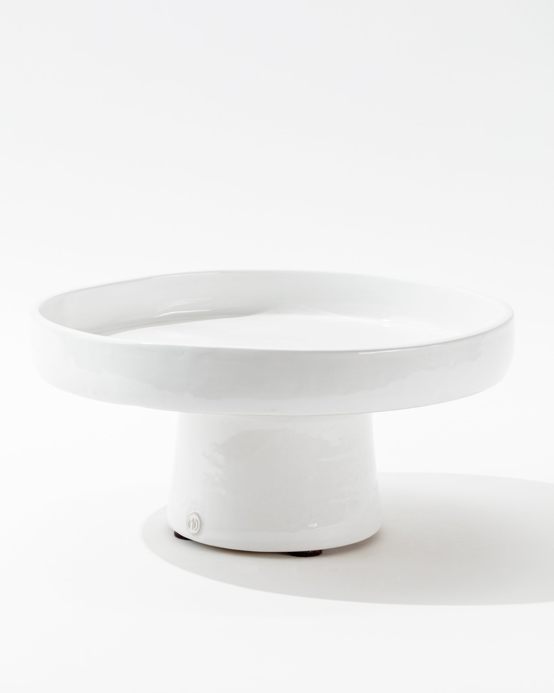 The 4807-L Cake Stand no. 807 L by Montes Doggett is an artisanal piece crafted from high-fired white ceramic. It showcases a round, flat surface supported by a wide, cylindrical base, and features a glossy finish with a minimalistic design against its white background.
