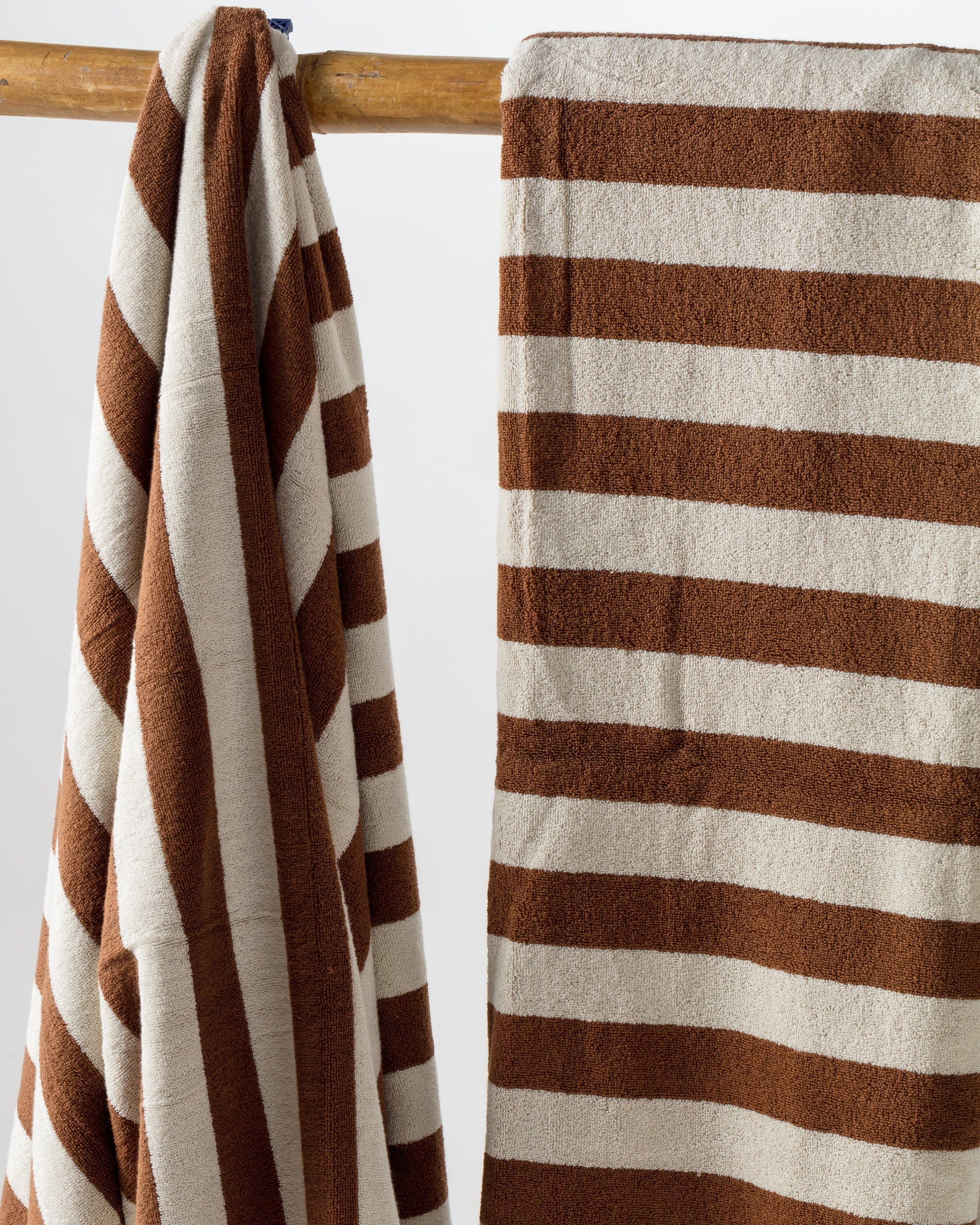 Two luxurious Maria Pool Towels by Autumn Sonata, featuring thick horizontal brown and white stripes, hang on a wooden rod. One towel is neatly folded over the rod, while the other is draped and slightly wrinkled. The background is plain and light-colored.