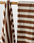 Two luxurious Maria Pool Towels by Autumn Sonata, featuring thick horizontal brown and white stripes, hang on a wooden rod. One towel is neatly folded over the rod, while the other is draped and slightly wrinkled. The background is plain and light-colored.