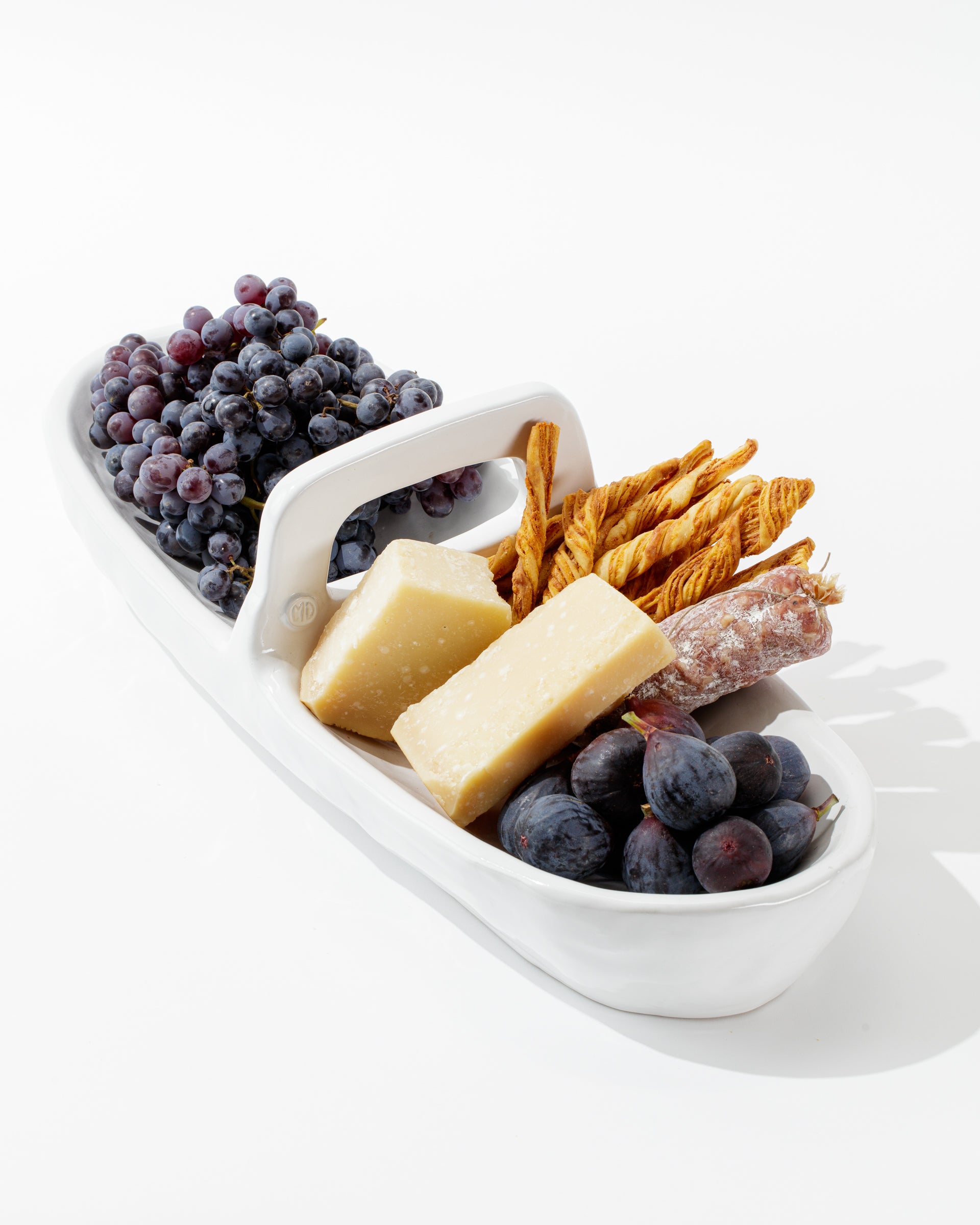 The 4774 Appetizer Platter NO. 774 by Montes Doggett elegantly holds a selection of grapes, two large pieces of cheese, breadsticks, and a piece of cured sausage. The food is artistically arranged against a white background, highlighting the platter&#39;s traditional craftsmanship and artisanal charm.