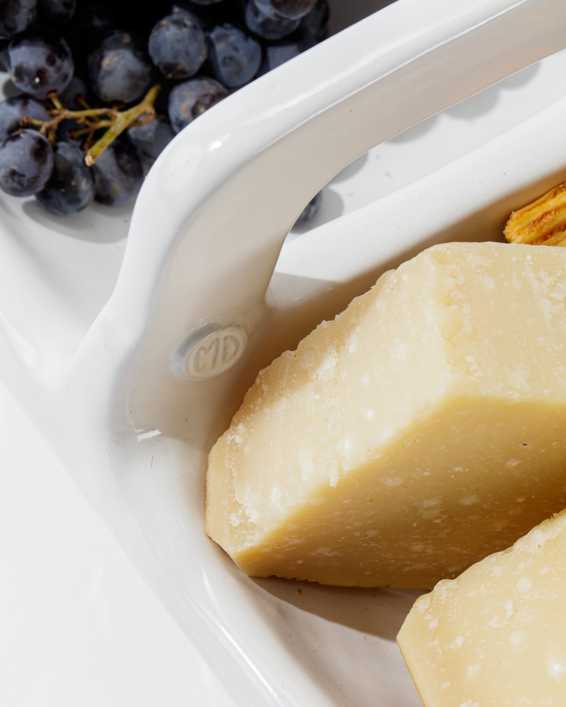 Close-up of two wedges of pale yellow cheese in a Montes Doggett 4774 Appetizer Platter NO. 774 handmade ceramic dish, with a cluster of dark grapes in the background. The image focuses on the texture of the cheese and the sleek design of the dish.