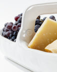 A close-up image of a white *handmade ceramic* basket from the 4774 Appetizer Platter NO. 774 collection by Montes Doggett, containing chunks of hard cheese and a bunch of grapes. The basket handle has the initials "MD" embossed on it, showcasing traditional techniques. The background is plain white.