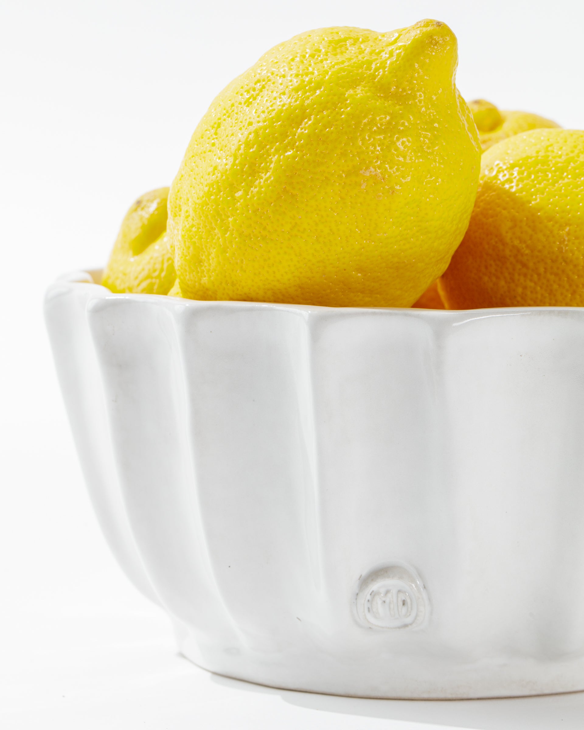 A white, high-fired ceramic 5131 bowl no. 131 by Montes Doggett, filled with bright yellow lemons. The artisanal bowl has a smooth, glossy texture with subtle vertical grooves and is handcrafted in Peru. The vibrant and fresh lemons are positioned to showcase their textured peels as the elegant bowl rests on a clean, white surface.