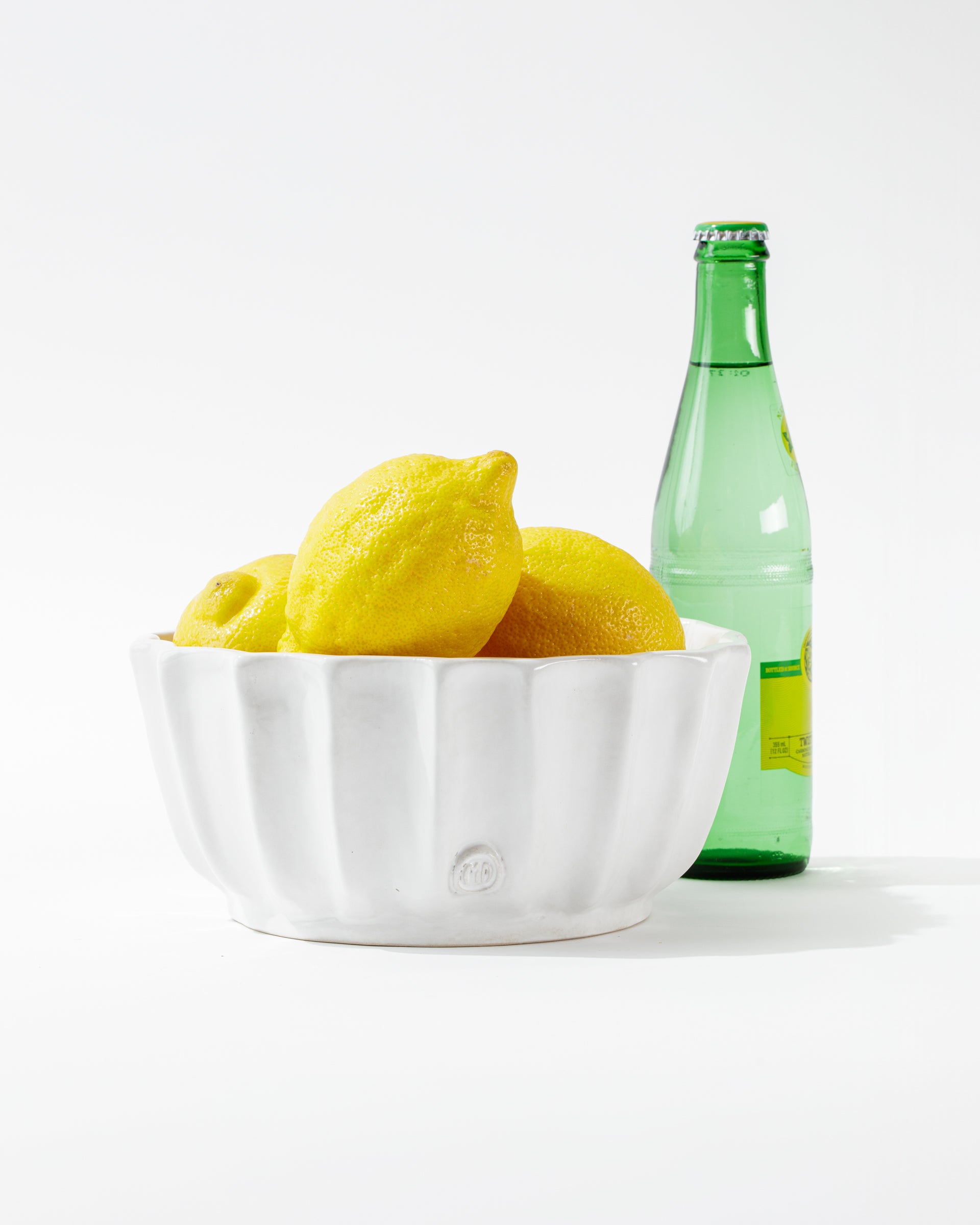 A white Montes Doggett 5131 Bowl No. 131 filled with bright yellow lemons is positioned in front of a green glass bottle of sparkling water, set against a clean white background.