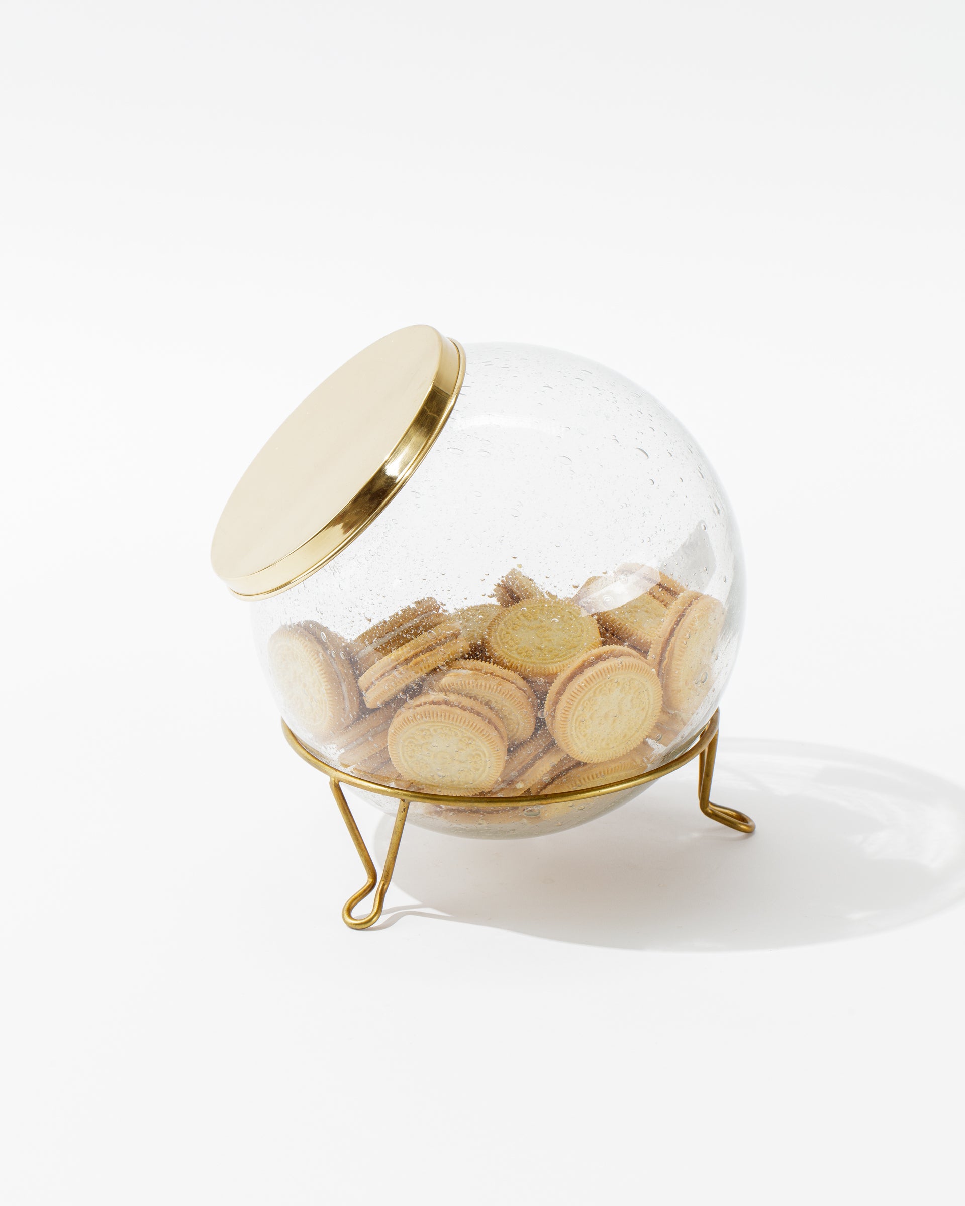 A Sir/Madam Scullery Jar, hand-blown from glass and topped with a polished brass lid, filled with vanilla sandwich cookies. The container is tilted on a three-legged gold stand, set against a white background.