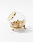 A Sir/Madam Scullery Jar, hand-blown from glass and topped with a polished brass lid, filled with vanilla sandwich cookies. The container is tilted on a three-legged gold stand, set against a white background.