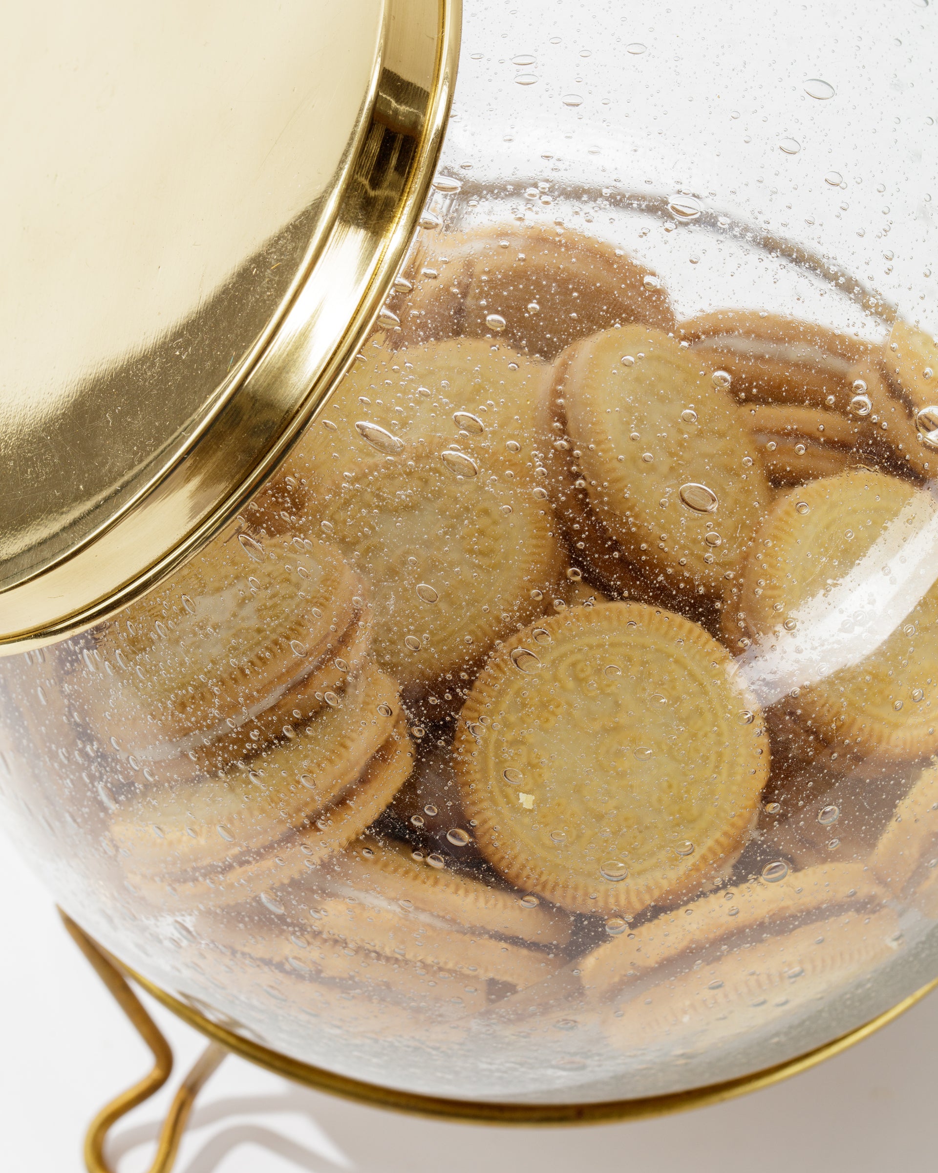 A clear, hand-blown Scullery Jar from Sir/Madam, featuring a polished brass lid and base, is filled with numerous round sandwich cookies. The jar has droplets of water on the inside, indicating condensation. The cookies appear to be vanilla-flavored or a similar light flavor.