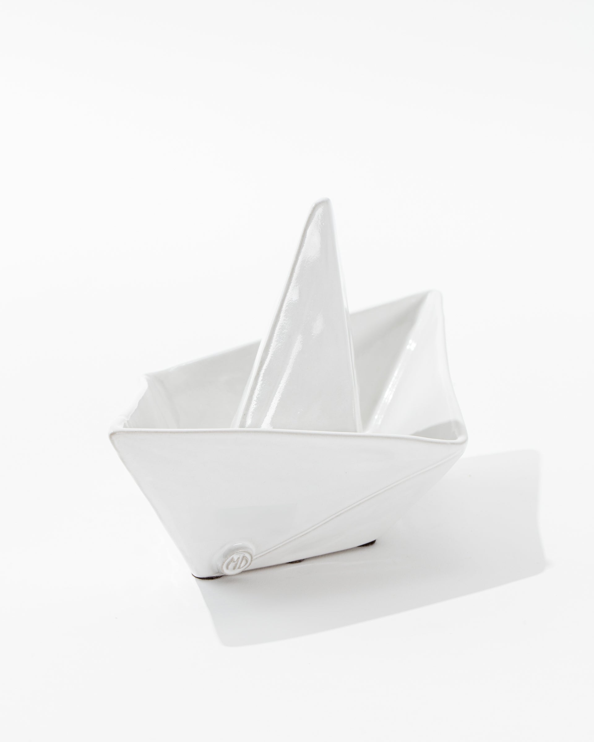 Introducing the 5419 Sail Boat no. 419 by Montes Doggett—a handcrafted white ceramic sculpture that beautifully captures the essence of high fired ceramic craftsmanship from Peru. This minimalist sailboat, with its clean, sharp lines and glossy finish, resembles an origami paper boat and is set against a plain white background.