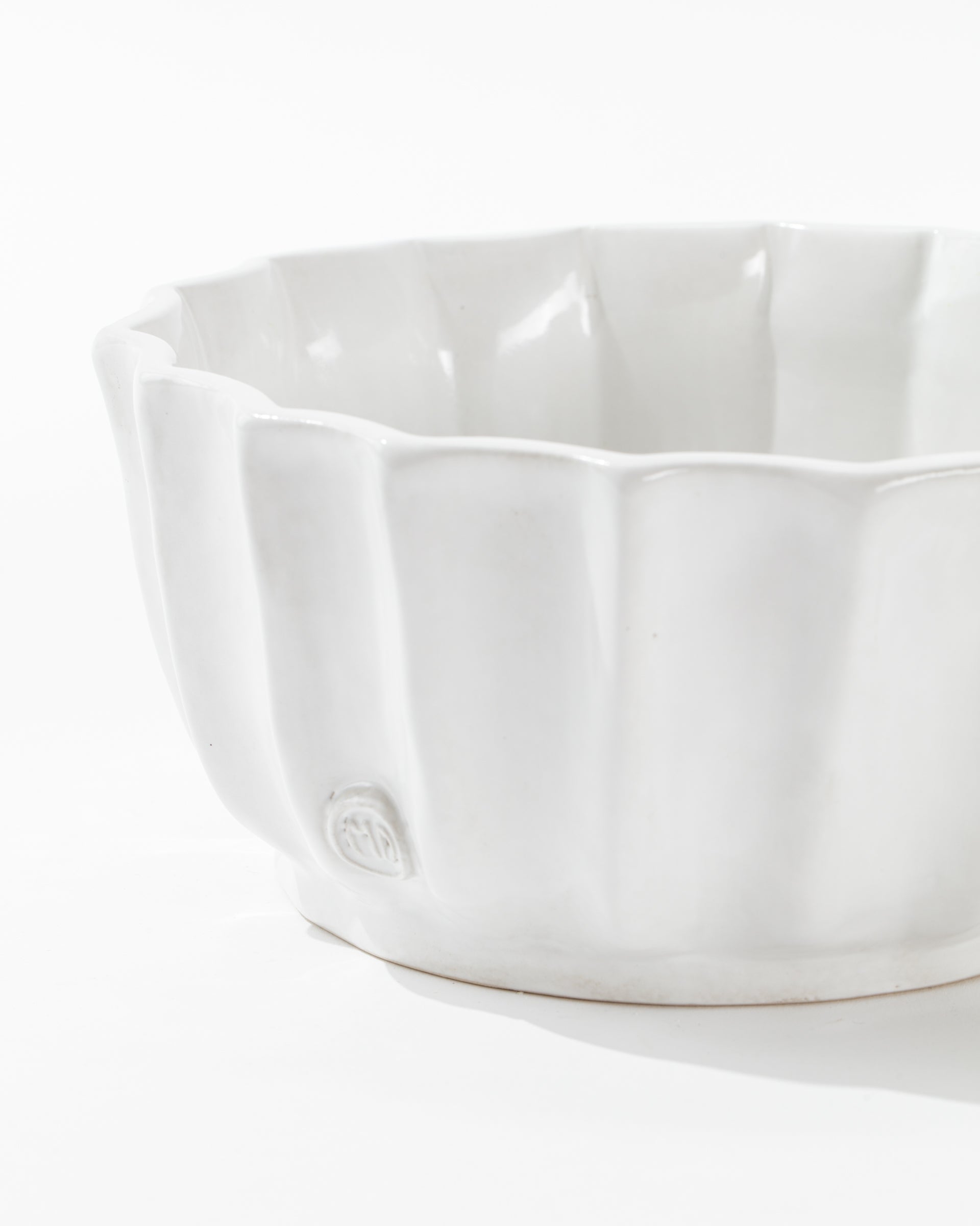 The 5131 bowl no. 131 by Montes Doggett is a white, high-fired ceramic bowl featuring smooth vertical ridges and a slight sheen. It includes a small emblem embossed on the lower part of the side near the base, highlighting artisanal craftsmanship. The backdrop is plain and white.