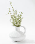 The 5431 Pitcher No. 431 by Montes Doggett, a white ceramic jug with a curved handle handmade in Peru, contains a bouquet of delicate green leafy stems. This high-fired jug features a smooth, glossy finish against a plain, bright white background, highlighting the simplicity and elegance of the arrangement.