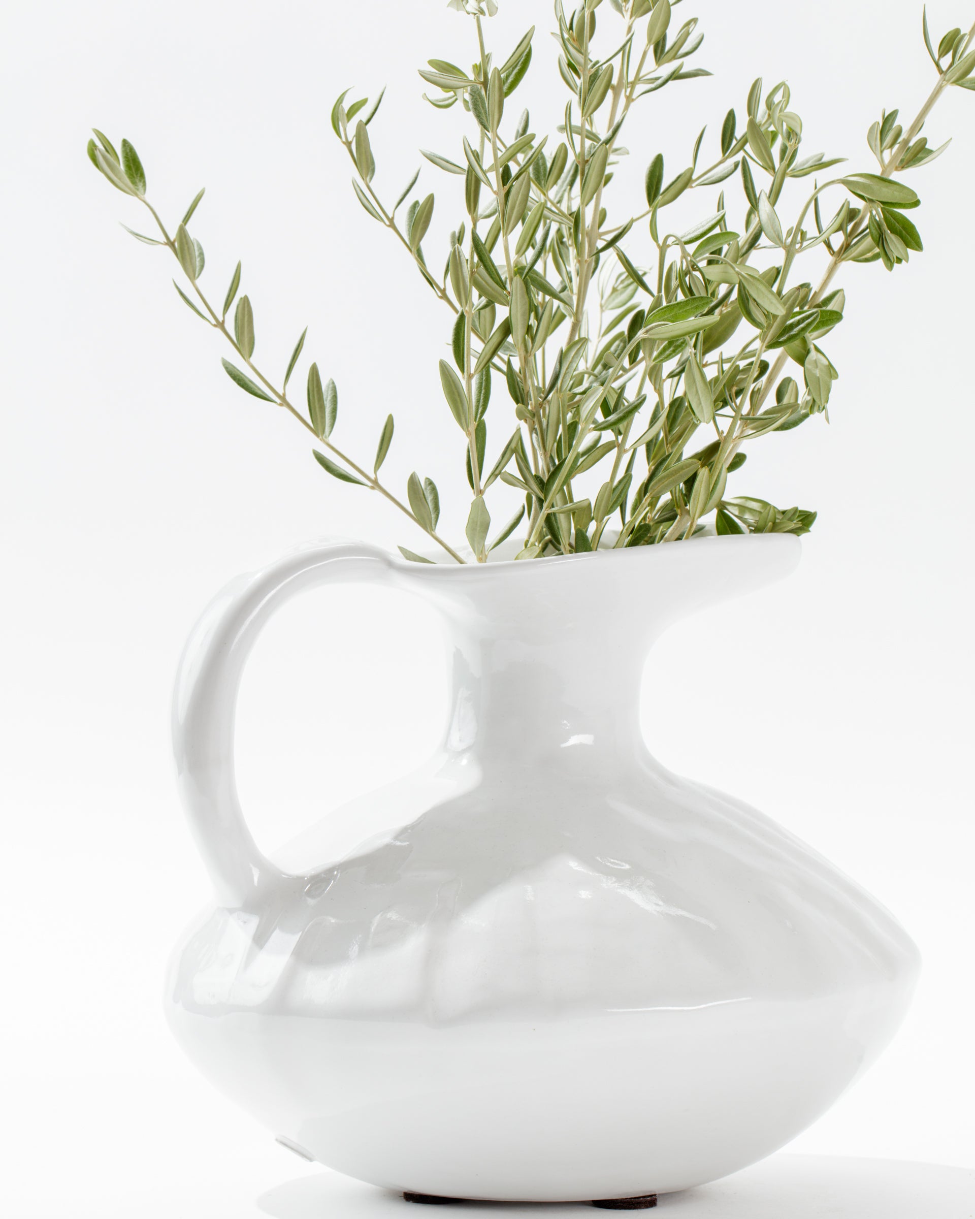 The 5431 Pitcher No. 431 by Montes Doggett is a white porcelain vase with a rounded body, a wide spout, and a handle. Handmade in Peru, this high-fired ceramic vase boasts a glossy finish and holds a bunch of green, leafy branches. The arrangement is displayed on a white surface against a white background.