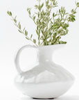 The 5431 Pitcher No. 431 by Montes Doggett is a white porcelain vase with a rounded body, a wide spout, and a handle. Handmade in Peru, this high-fired ceramic vase boasts a glossy finish and holds a bunch of green, leafy branches. The arrangement is displayed on a white surface against a white background.