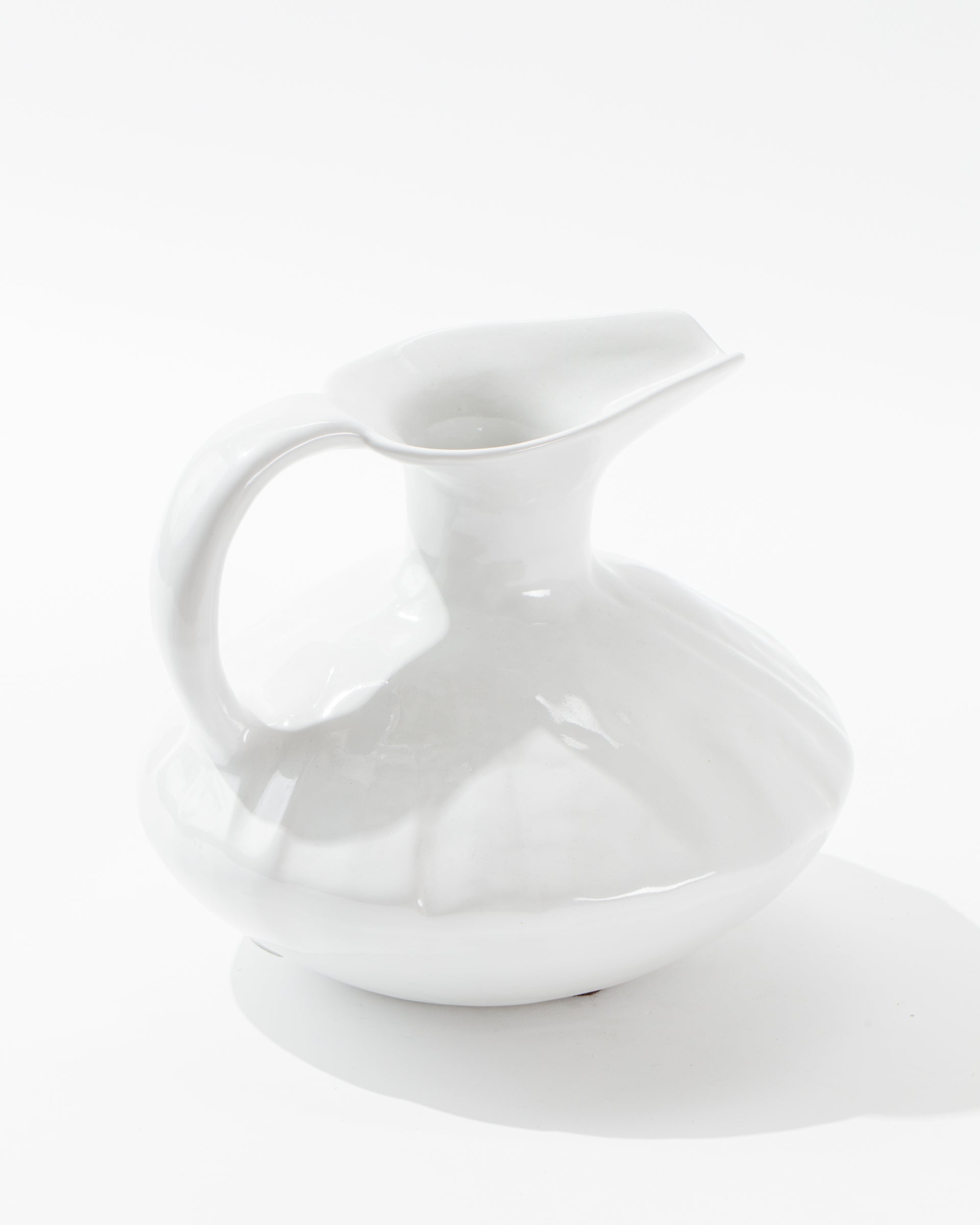 The Montes Doggett 5431 Pitcher No. 431, a glossy white ceramic pitcher handmade in Peru, features a large rounded base, a curved handle, and a wide spouted opening. Its high-fired design presents a sleek and minimalistic appearance against a plain white background.