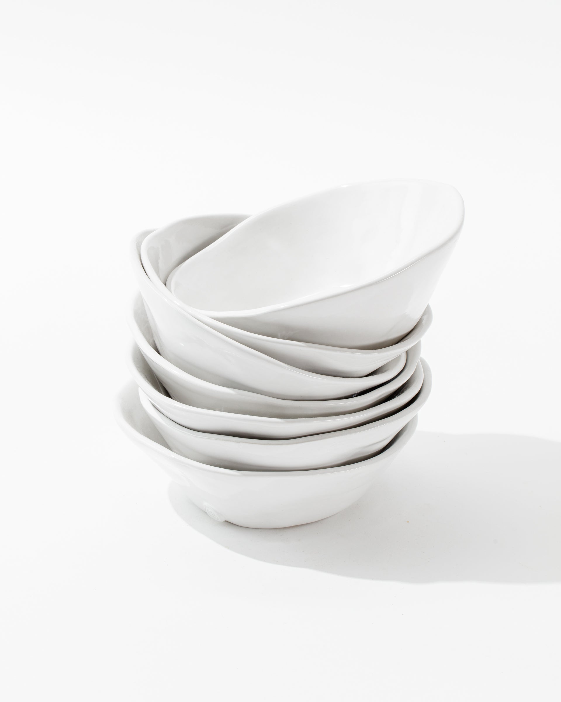 A set of six 5204-L Bowl No. 204 L white, oval-shaped ceramic bowls from Montes Doggett, handmade by Peruvian artisans, is arranged in an overlapping manner against a plain white background. These durable dinnerware pieces feature smooth surfaces and a minimalist design, creating a clean and simple aesthetic.