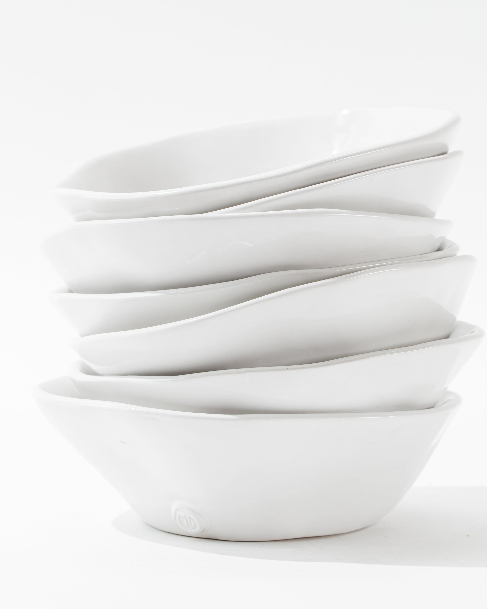 A stack of six 5204-L Bowl No. 204 L ceramic bowls from Montes Doggett, white and irregularly shaped, handcrafted by Peruvian artisans, arranged on a white surface against a white background. The bowls are nested unevenly, creating a wavy and dynamic appearance that showcases durable dinnerware with artisanal charm.