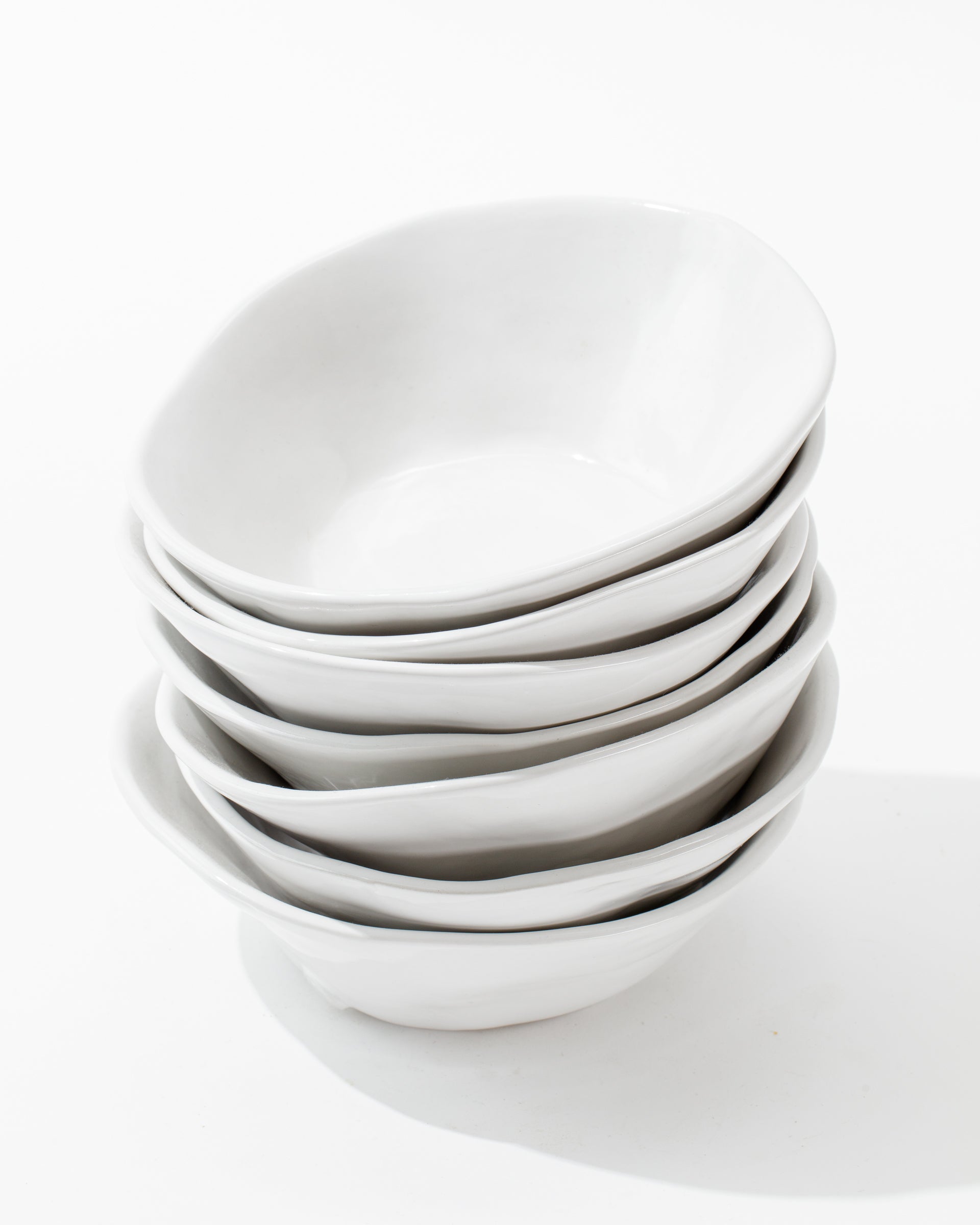 A stack of six 5204-L Bowl No. 204 L white porcelain bowls with slightly uneven, organic edges, crafted by Peruvian artisans from Montes Doggett, is placed against a plain white background. The bowls are neatly arranged, one on top of the other, displaying a handmade ceramic bowl&#39;s minimalist and clean aesthetic.