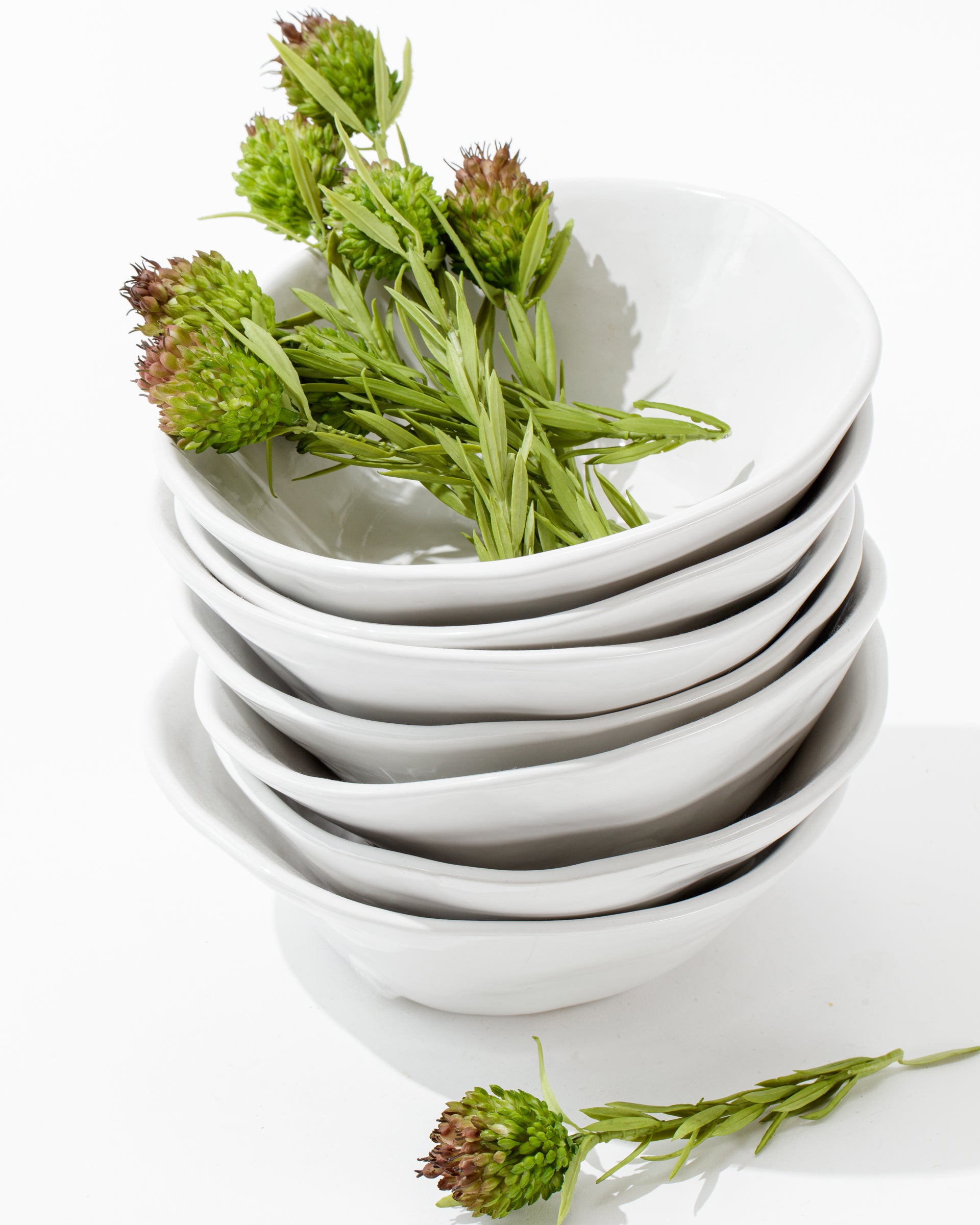 A stack of Montes Doggett 5204-L Bowl No. 204 L high-fired ceramic bowls with a few green and brown spiky flowers on top and one beside them sits on the white surface. The handmade ceramic bowls and flowers are arranged against a plain white background, showcasing an elegant simplicity.