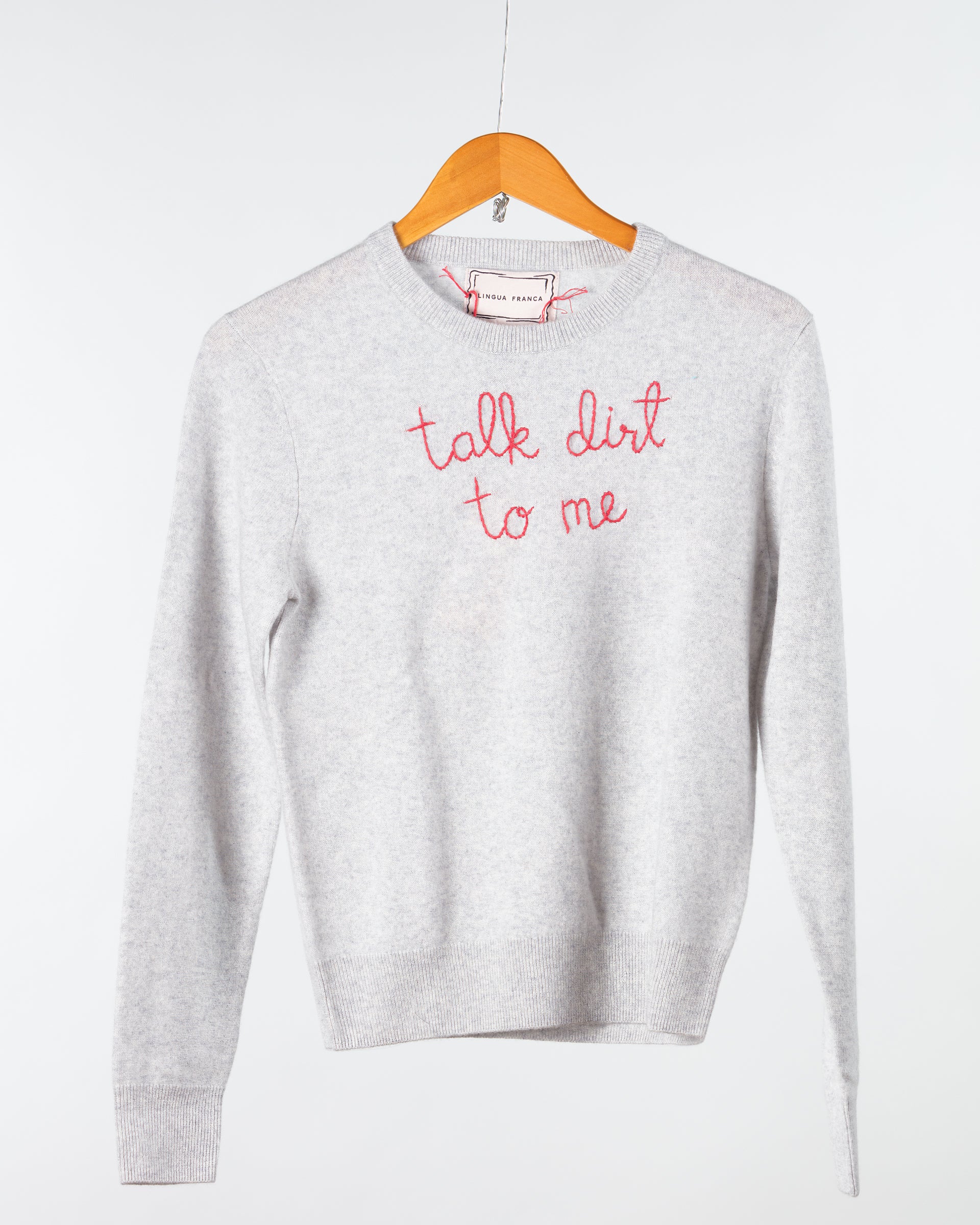 A light gray Lingua Franca Cashmere Crewneck sweater with "talk dirt to me" embroidered in pinkish-red hangs on a wooden hanger against a plain background.