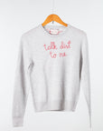 A light gray Lingua Franca Cashmere Crewneck sweater with "talk dirt to me" embroidered in pinkish-red hangs on a wooden hanger against a plain background.