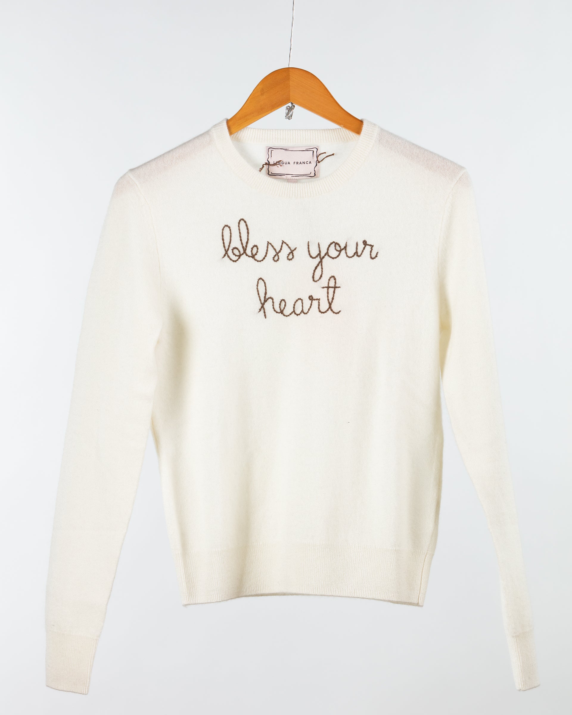 A cream Lingua Franca Cashmere Crewneck elegantly hangs on a wooden hanger, featuring "bless your heart" beautifully embroidered in cursive. Crafted from sustainably sourced materials, it rests against a white background, radiating timeless elegance.