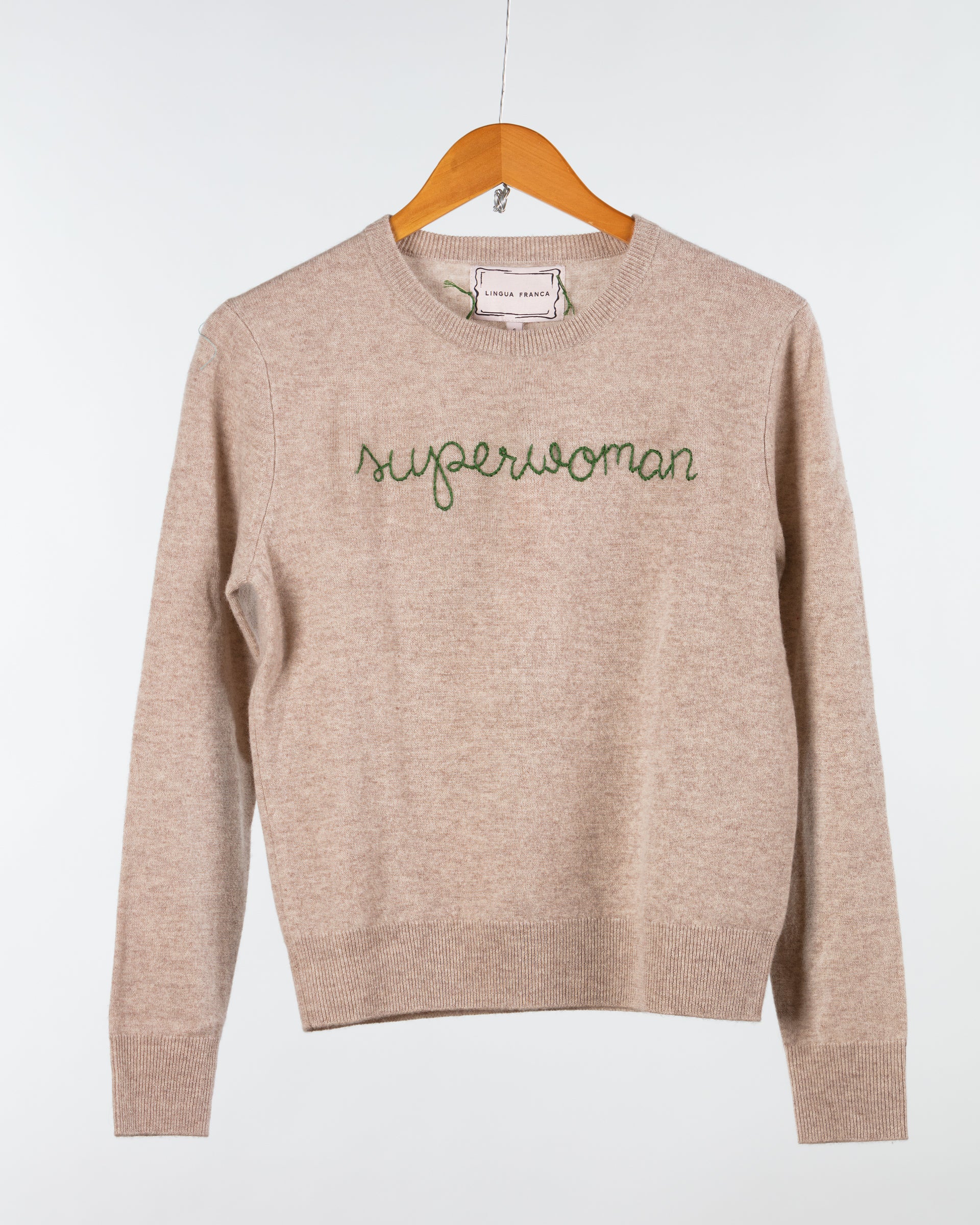 A beige Lingua Franca Cashmere Crewneck hangs on a wooden hanger against a plain backdrop. "Superwoman" is embroidered in green cursive on the front, showcasing its ethical production.