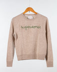 A beige Lingua Franca Cashmere Crewneck hangs on a wooden hanger against a plain backdrop. "Superwoman" is embroidered in green cursive on the front, showcasing its ethical production.