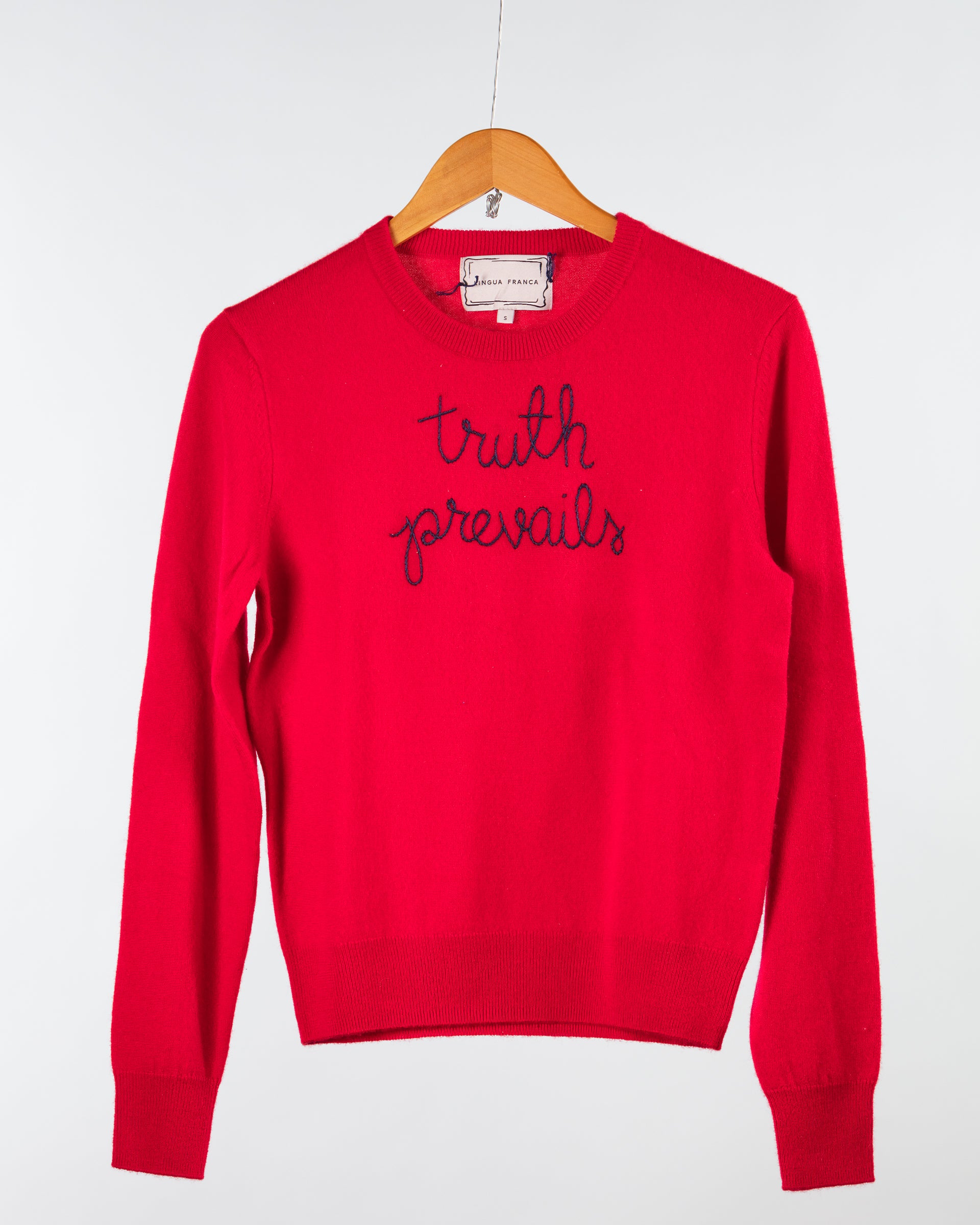 A bright red Cashmere Crewneck by Lingua Franca hangs on a wooden hanger, featuring "truth prevails" embroidered in black script across the chest. Ethically crafted against a plain white backdrop, this stylish piece is as mindful as it is chic.