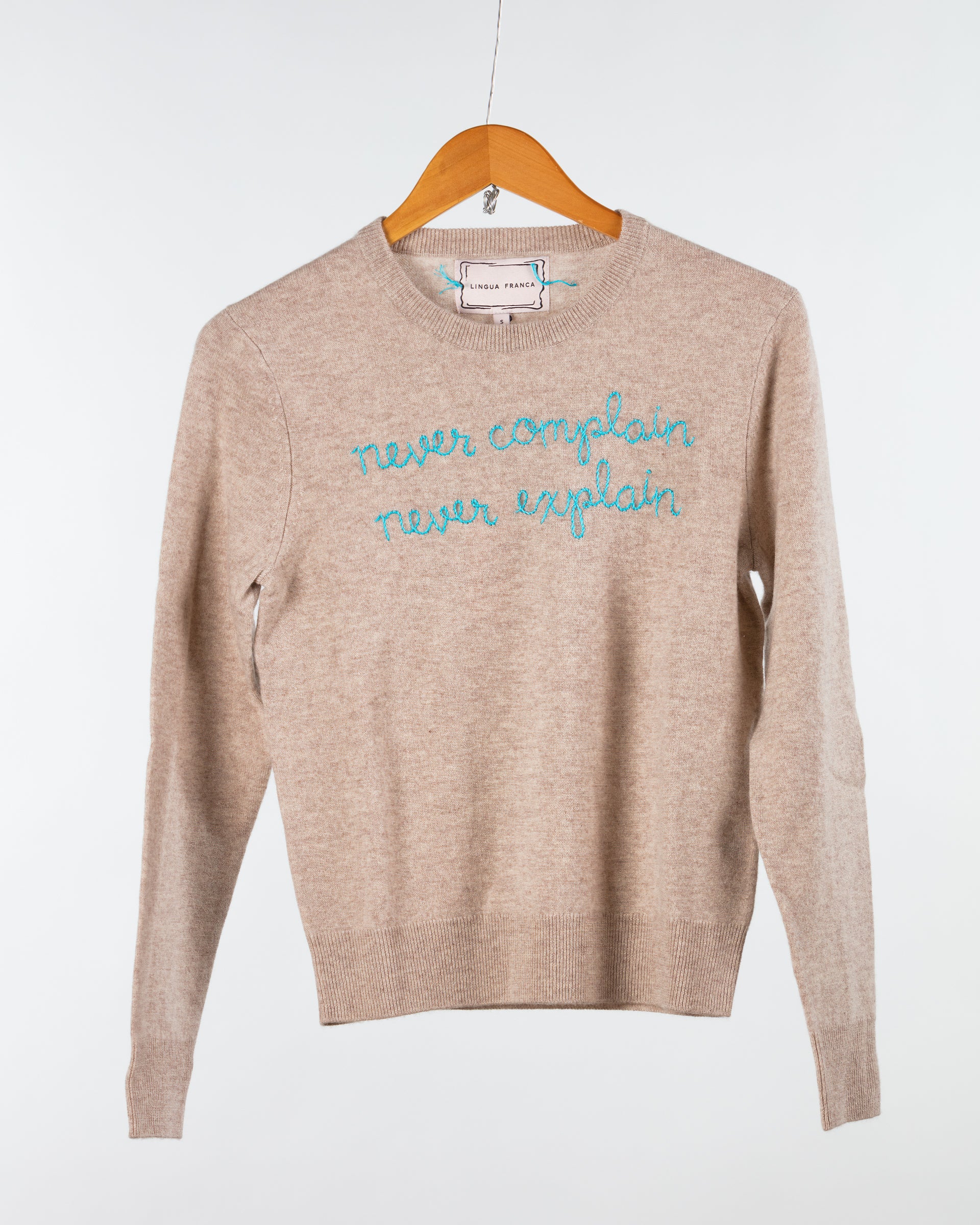 The Lingua Franca Cashmere Crewneck is a beige sweater on a wooden hanger, adorned with blue text stating "never complain, never explain." Stylish and ethically made, it reflects quality and sustainability.