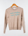 The Lingua Franca Cashmere Crewneck is a beige sweater on a wooden hanger, adorned with blue text stating "never complain, never explain." Stylish and ethically made, it reflects quality and sustainability.