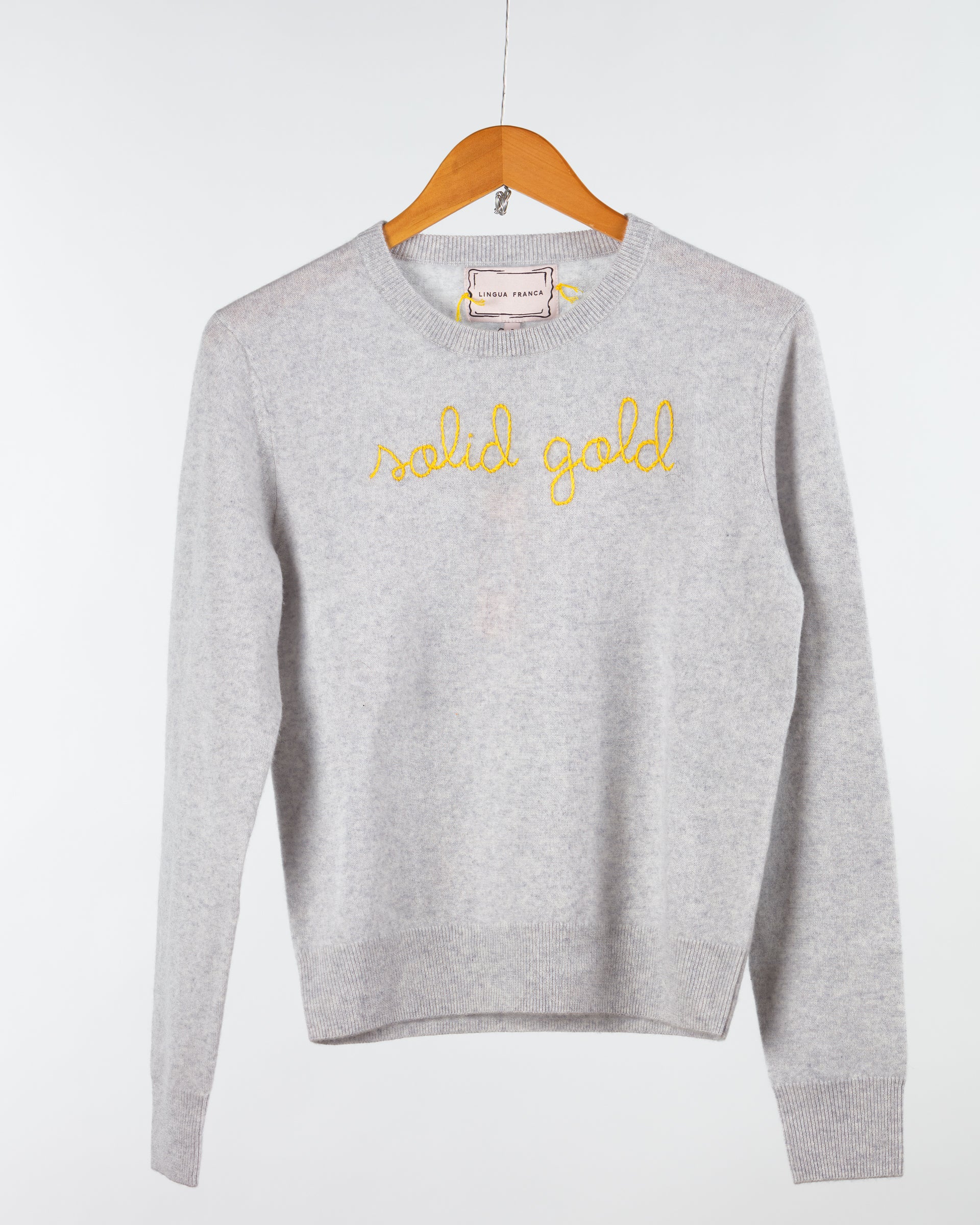 The Lingua Franca Cashmere Crewneck, embroidered with "solid gold" in yellow on the front, is displayed on a wooden hanger against a white background. This ethically crafted piece merges style with sustainability.