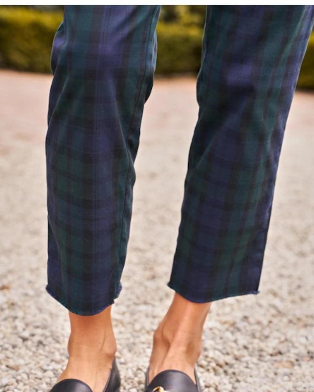 A person stands on gravel wearing Frank & Eileen's WICKLOW The Italian Chino in Italian Performance Twill featuring four-way stretch, paired with black loafers. Blurred greenery forms the background.