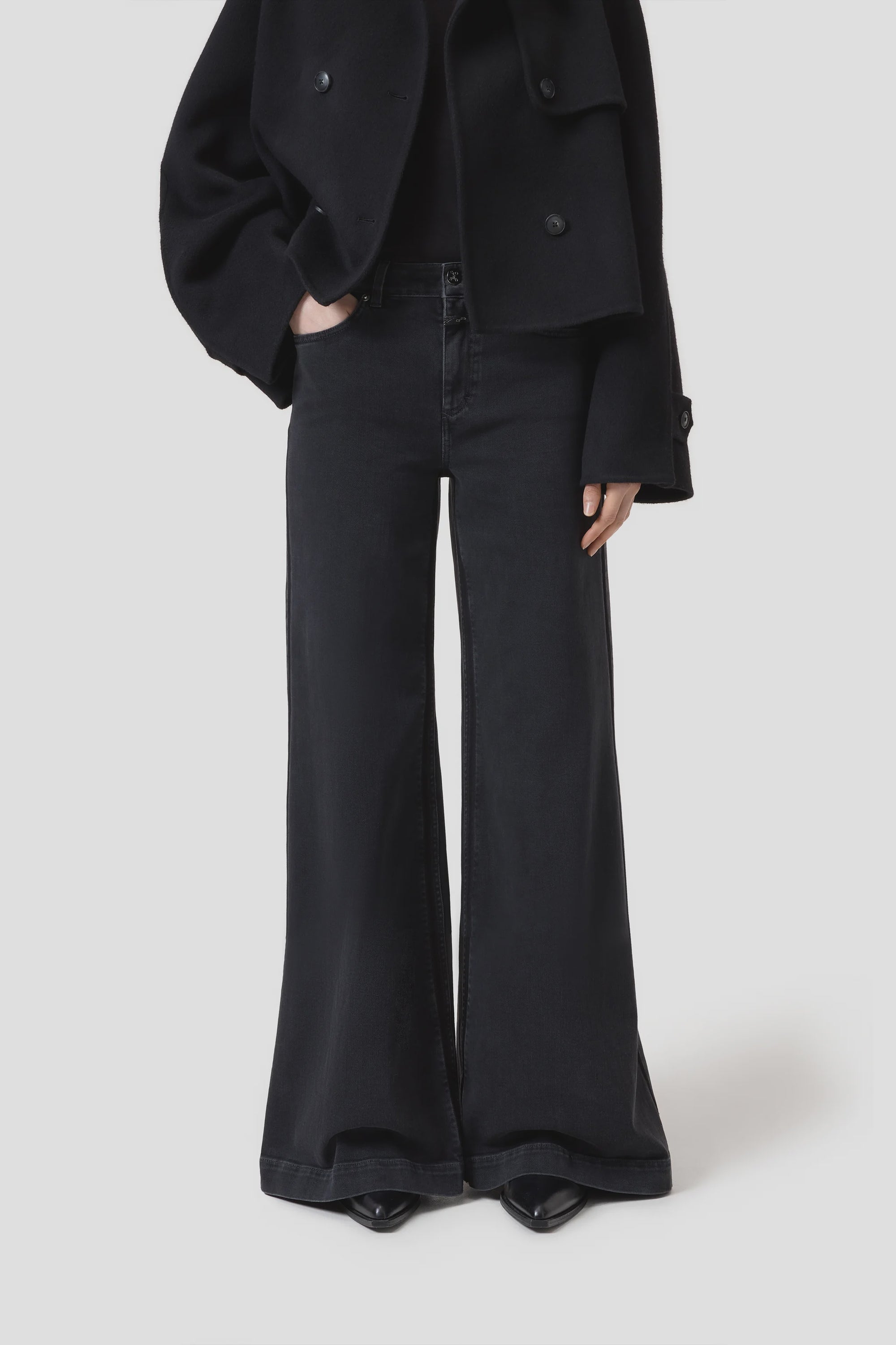 A person wears CLOSED's Glow Up black high waist flared leg jeans, a black coat, and black pointed-toe shoes, standing with one hand in their pocket against a plain light background. The upper part of the face is not visible.