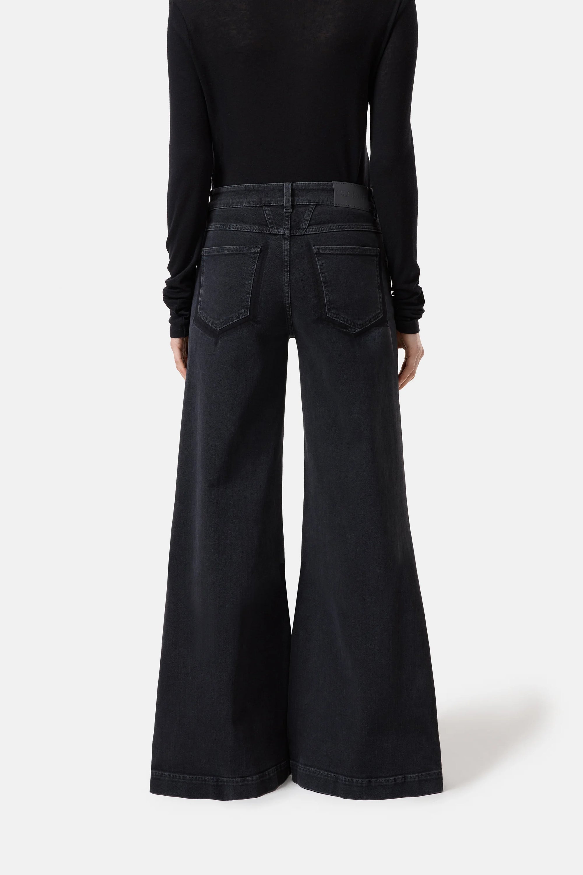 A person faces away against a white background, wearing CLOSED's Glow Up high-waisted black denim jeans with flared legs and large back pockets, paired with a black long-sleeve top.