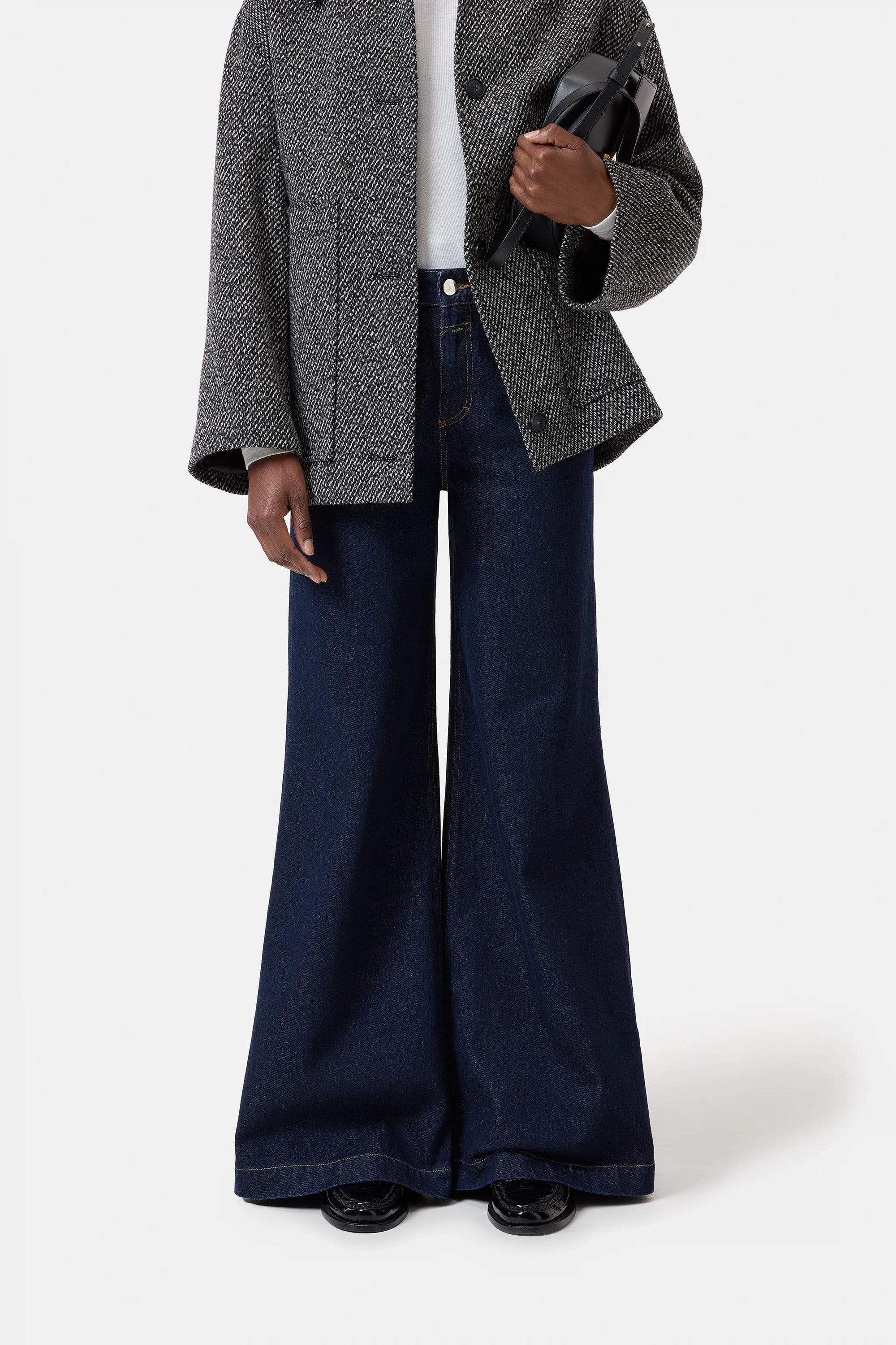 A person models the CLOSED "Glow Up" high waist flared leg dark blue jeans, styled with a textured gray jacket and black handbag, against a plain white background. The individual’s head is not visible in the image.