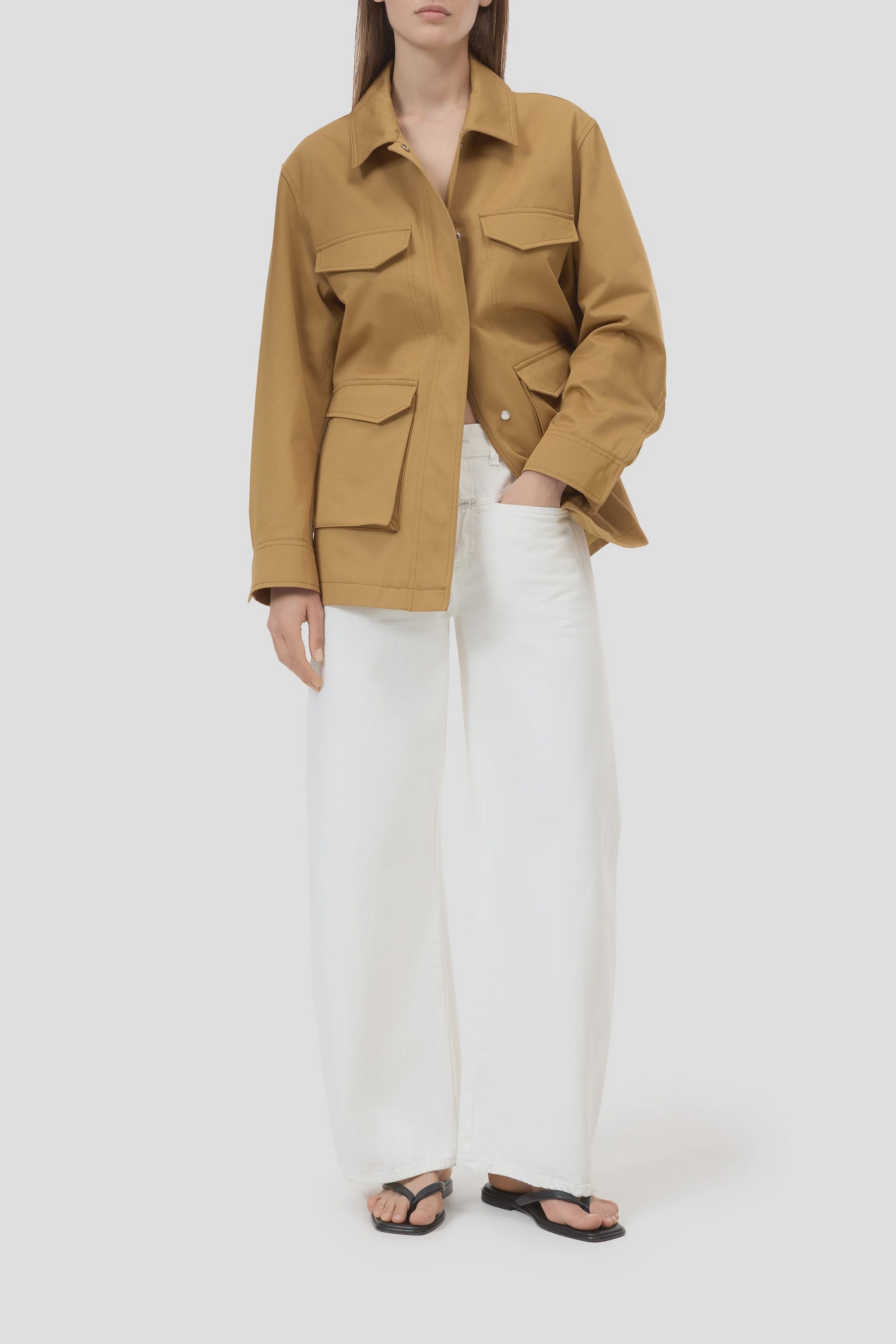 A person in CLOSED's Tori X Denim barrel-leg jeans and a tan utility jacket with large pockets, paired with black sandals, stands against a plain light gray background.