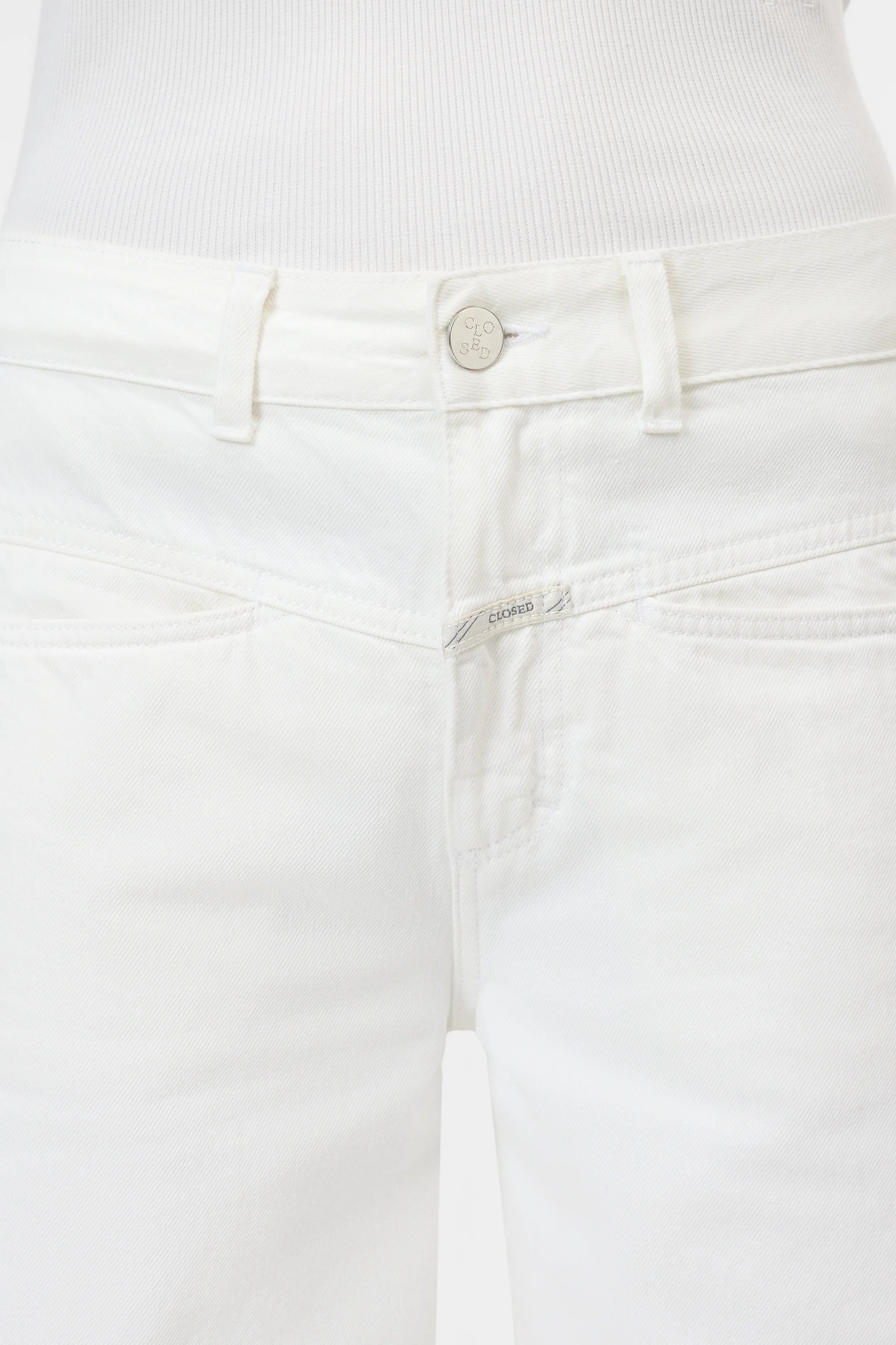 Close-up of a person wearing CLOSED Tori X Denim mid-rise white jeans, crafted from recycled cotton with a silver button, front pockets, subtle stitching, and a tucked-in white ribbed top.