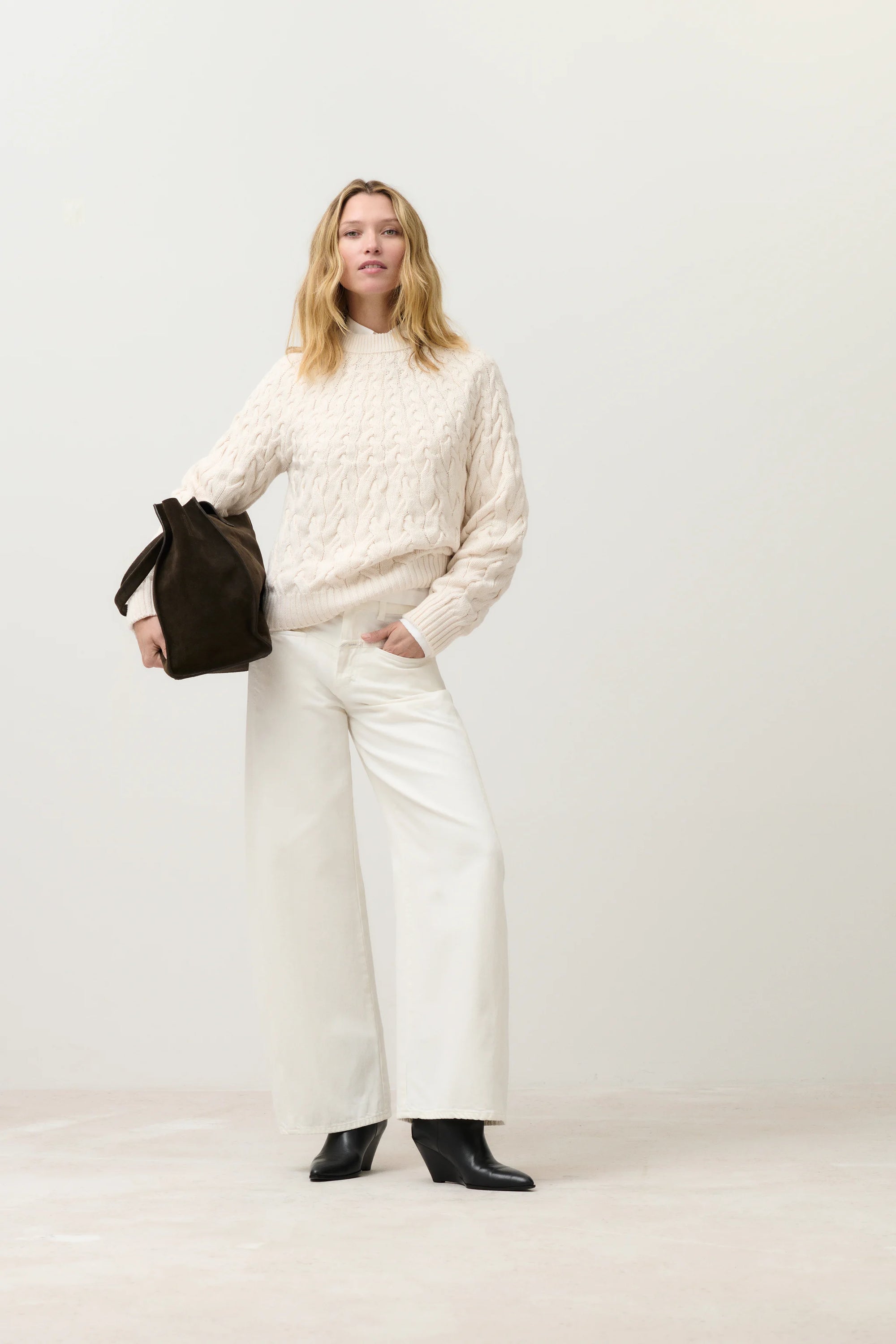 A woman stands against a plain background wearing CLOSED's Tori X Denim wide-leg pants, a cream recycled cotton cable-knit sweater, black ankle boots, and holding a large dark brown bag.