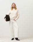 A woman stands against a plain background wearing CLOSED's Tori X Denim wide-leg pants, a cream recycled cotton cable-knit sweater, black ankle boots, and holding a large dark brown bag.