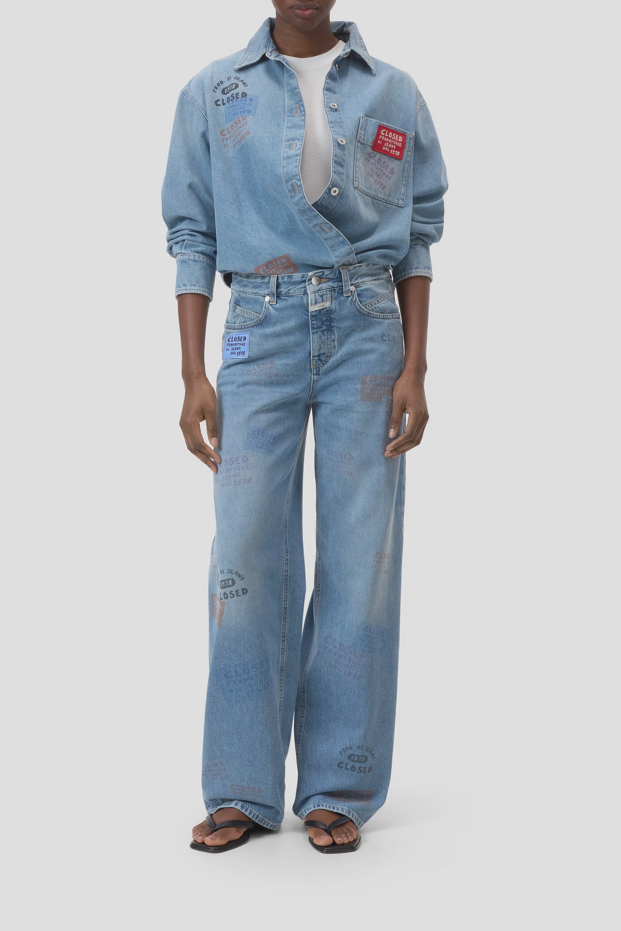 A person wears CLOSED’s Nikka Denim—a light blue eco-conscious shirt and matching wide-leg jeans with colored patches and text print—styled with a white top and black flip-flops, standing against a plain background.