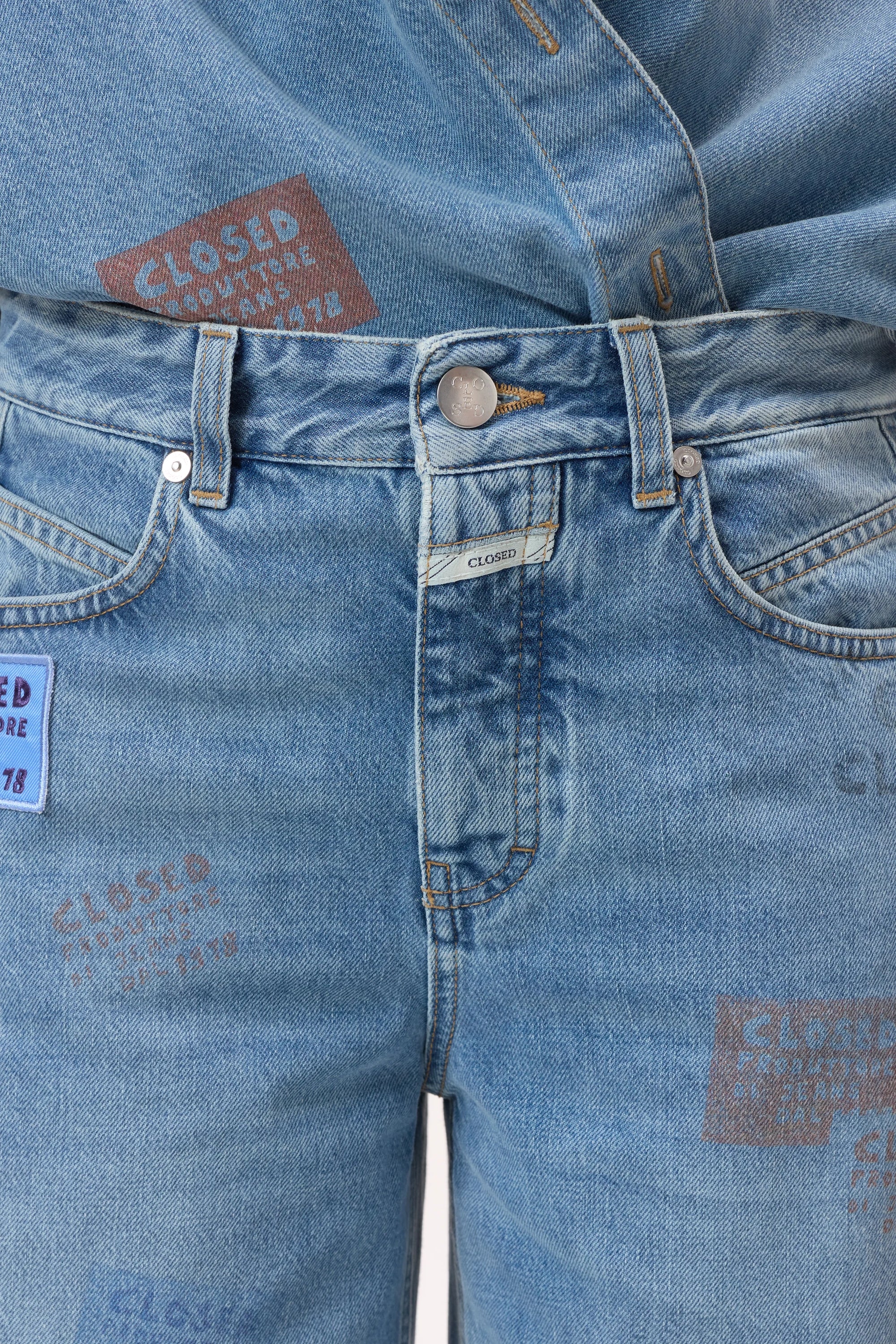 Close-up of a person wearing CLOSED's Nikka Denim jeans and matching shirt in light blue salt-and-pepper wash, featuring colorful "CLOSED" text patches and crafted with eco-conscious materials.