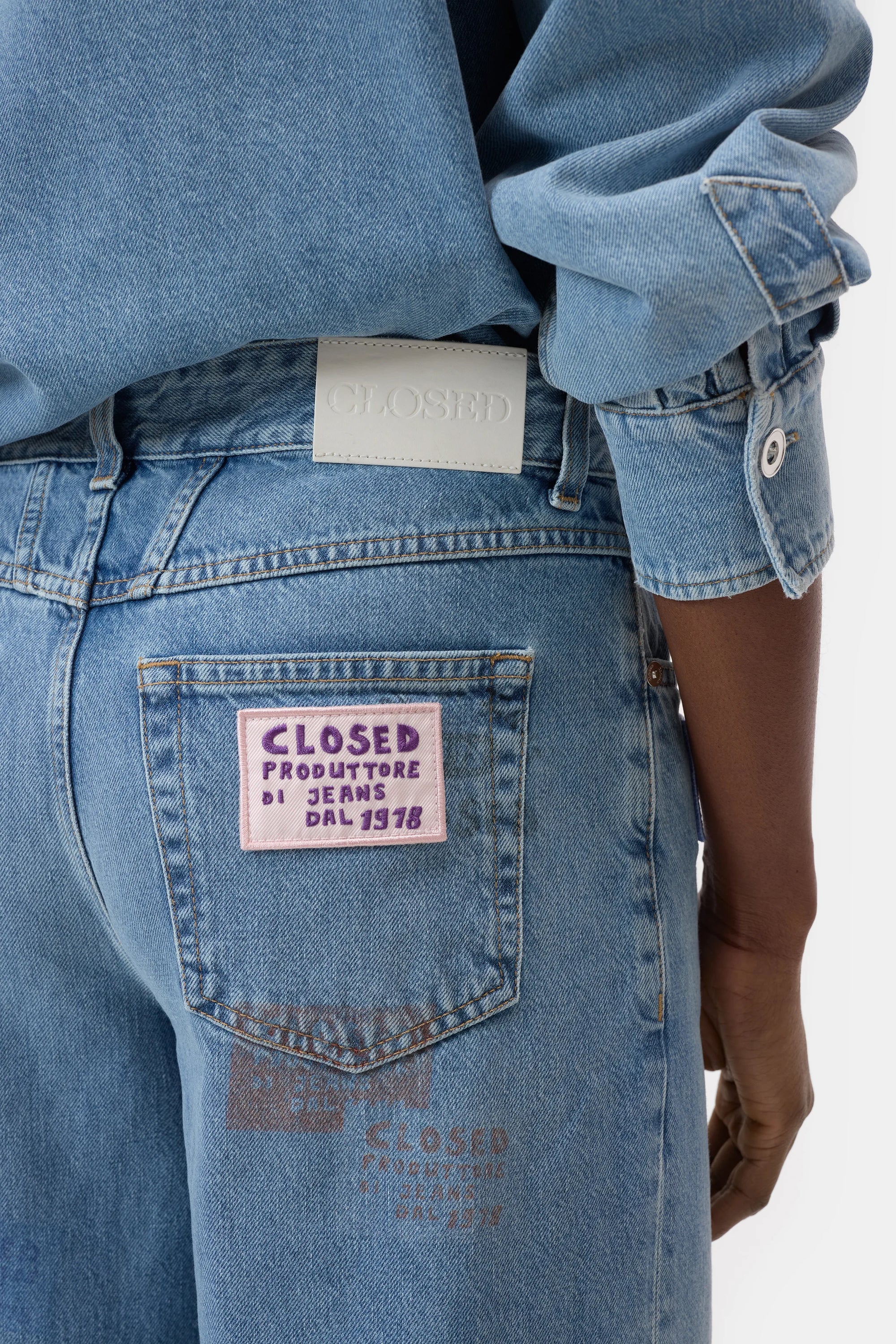 Someone wears light blue CLOSED Nikka Denim jeans and a matching shirt, highlighting the back pocket’s purple patch reading “CLOSED PRODUTTORE DI JEANS DAL 1978,” with a white label on the waistband.