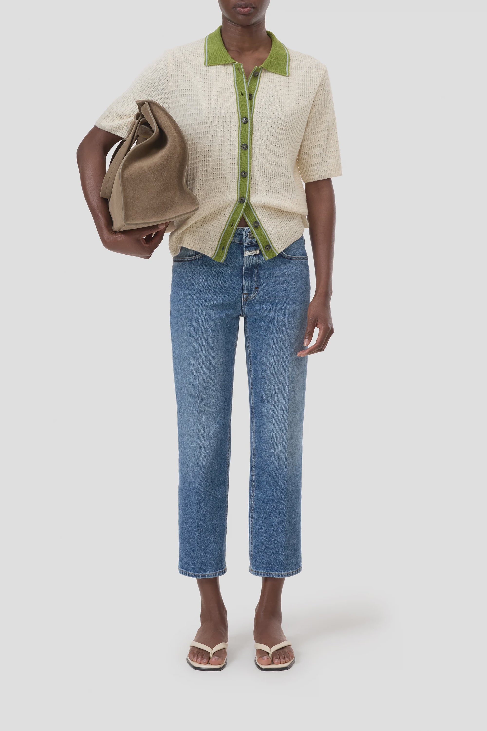 A person wearing CLOSED's Milo cream short-sleeve shirt with green trim, blue straight-leg jeans, white sandals, and carrying a large brown tote bag under their arm stands against a plain white background.