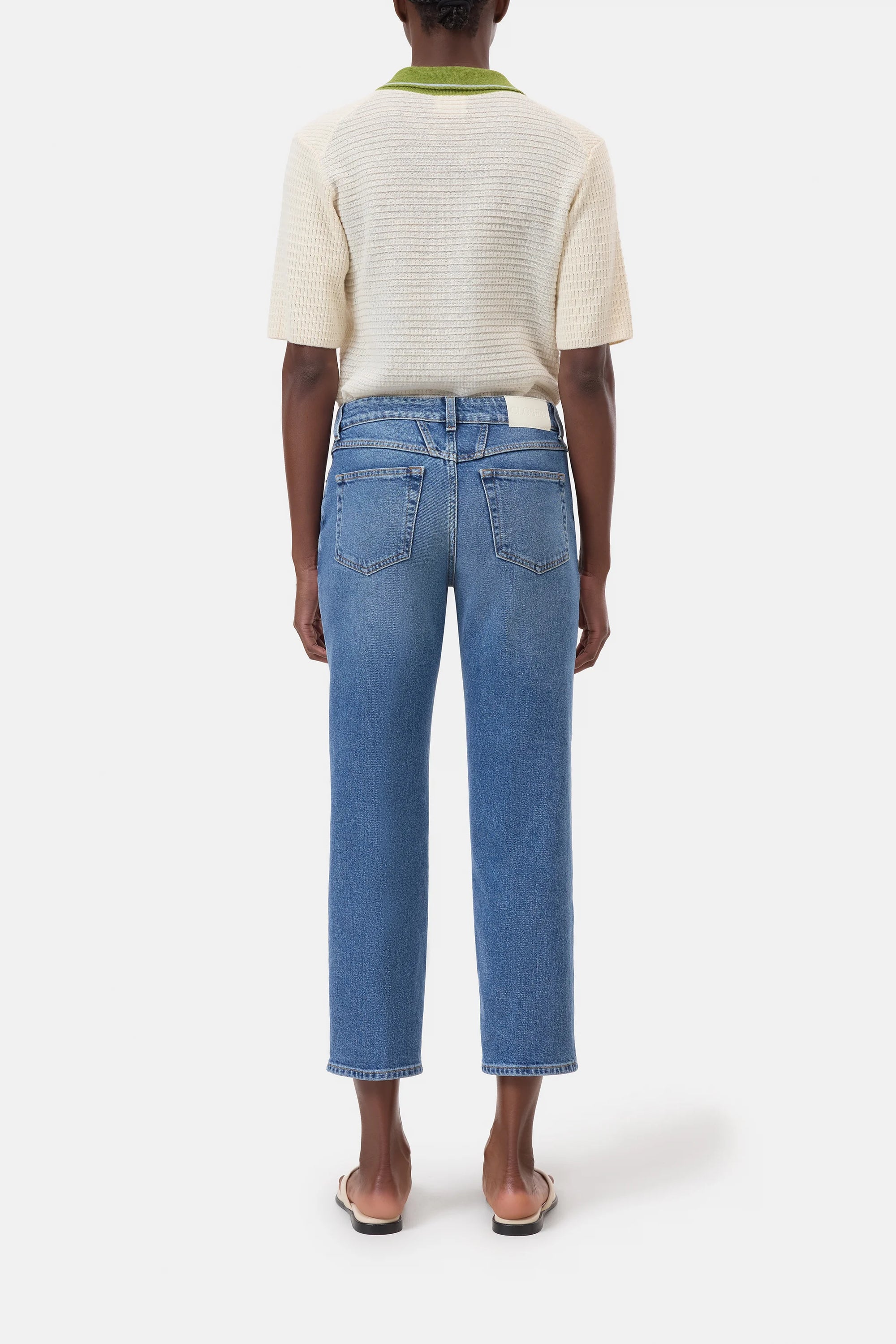 A person faces away, wearing CLOSED's Milo— a short-sleeved textured cream top with green trim, blue straight-leg sustainable denim jeans, and beige flat sandals—against a plain white background.